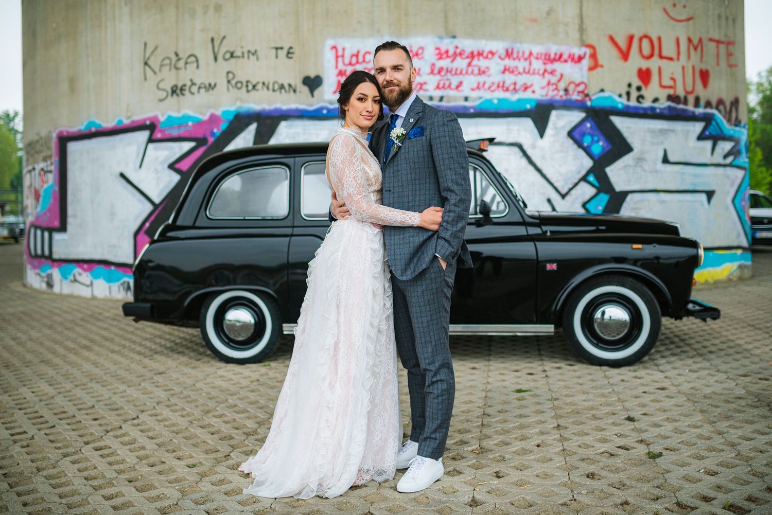 Bride and groom posing together in front of black vintage car and graffiti wall on wedding day.