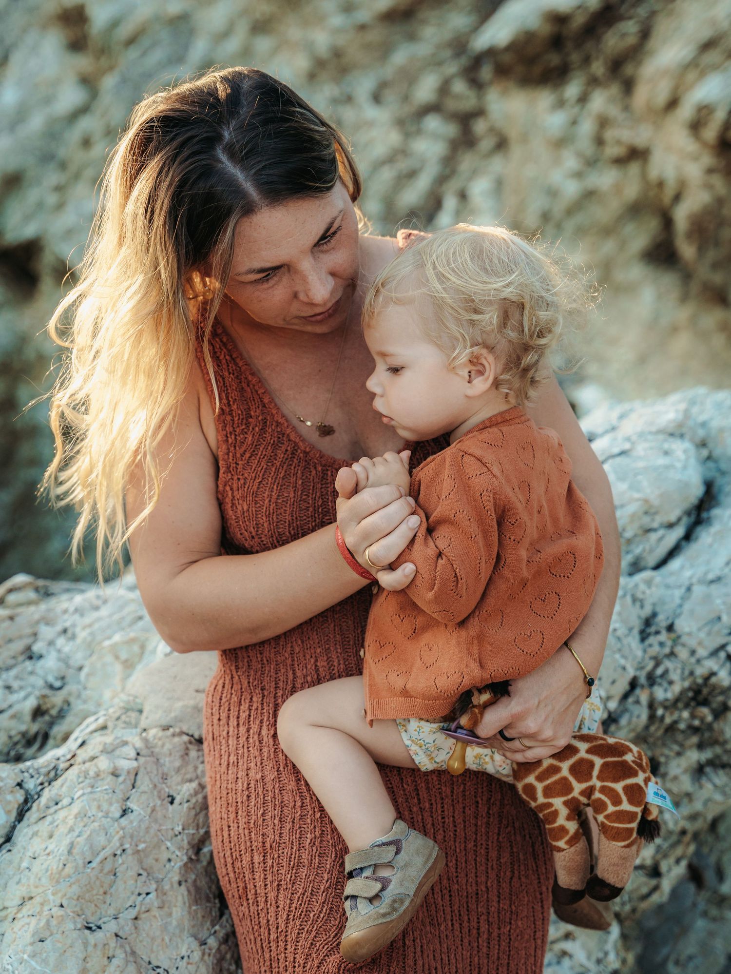 A woman in rust knit dress tenderly holds a curly-haired toddler clutching a giraffe toy on rocks.