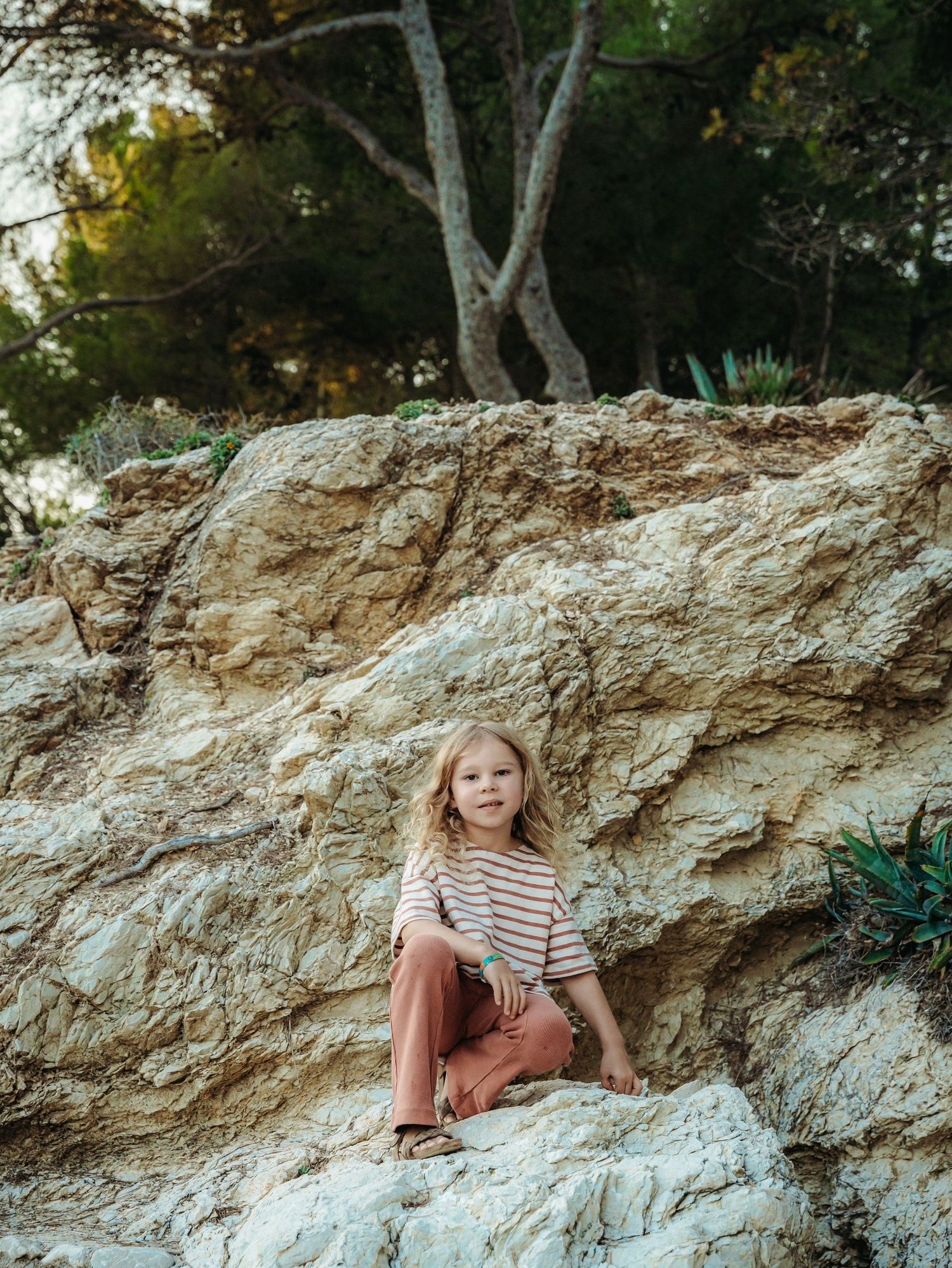 Blonde child in striped top and rust pants sits on large rocks surrounded by trees and lush greenery.