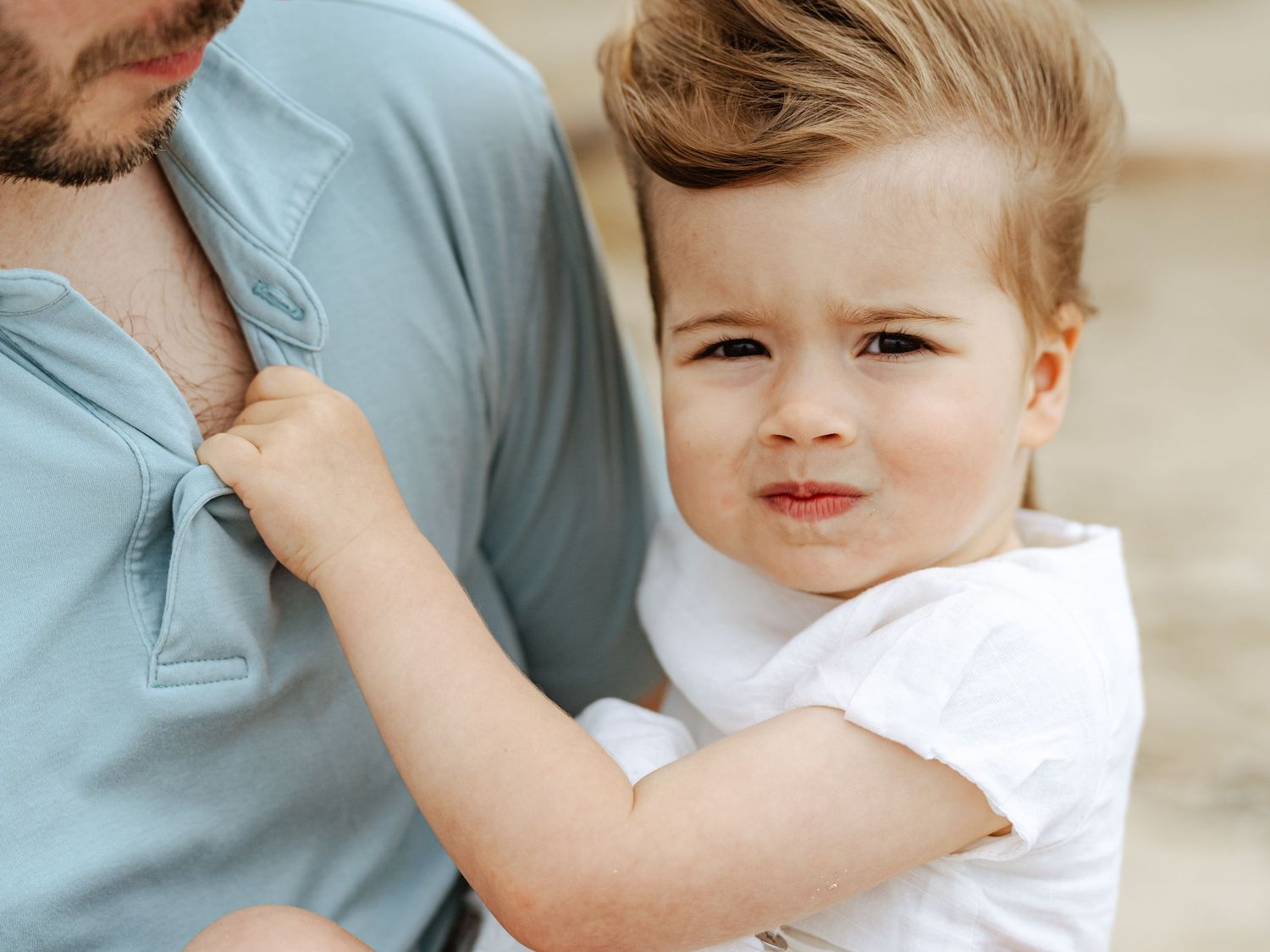 Toddler boy in white shirt clinging to adult in light blue polo during outdoor family photo session.