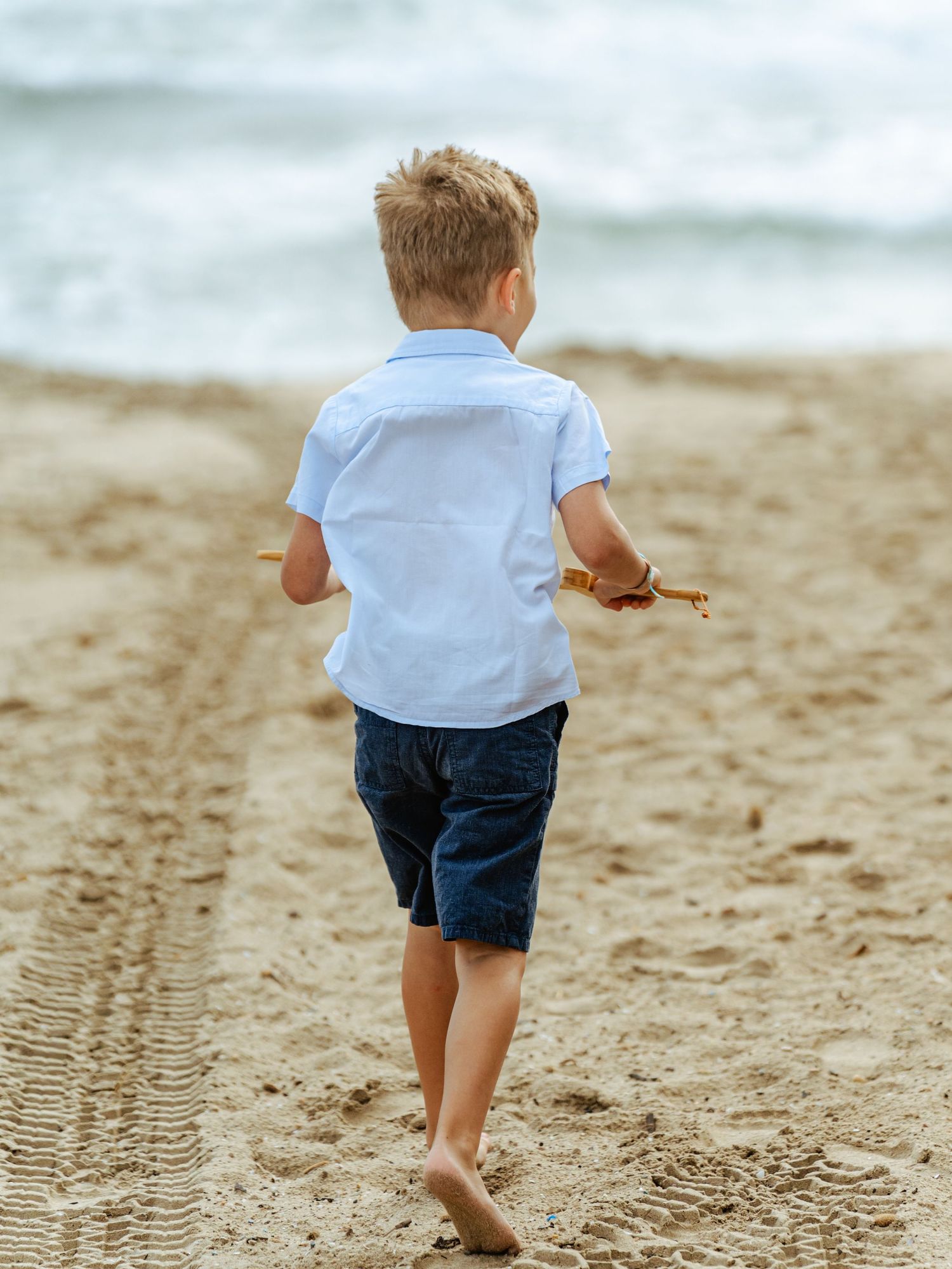 Young blonde boy in white shirt and blue shorts walking barefoot toward ocean on sandy beach.
