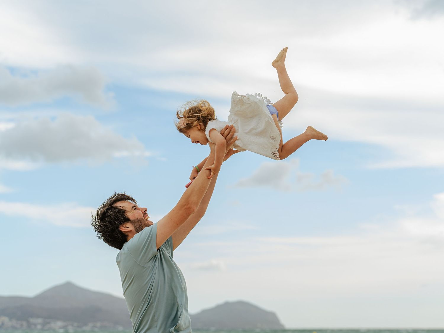 Father lifting child in white dress high into blue cloudy sky with mountains visible in background.