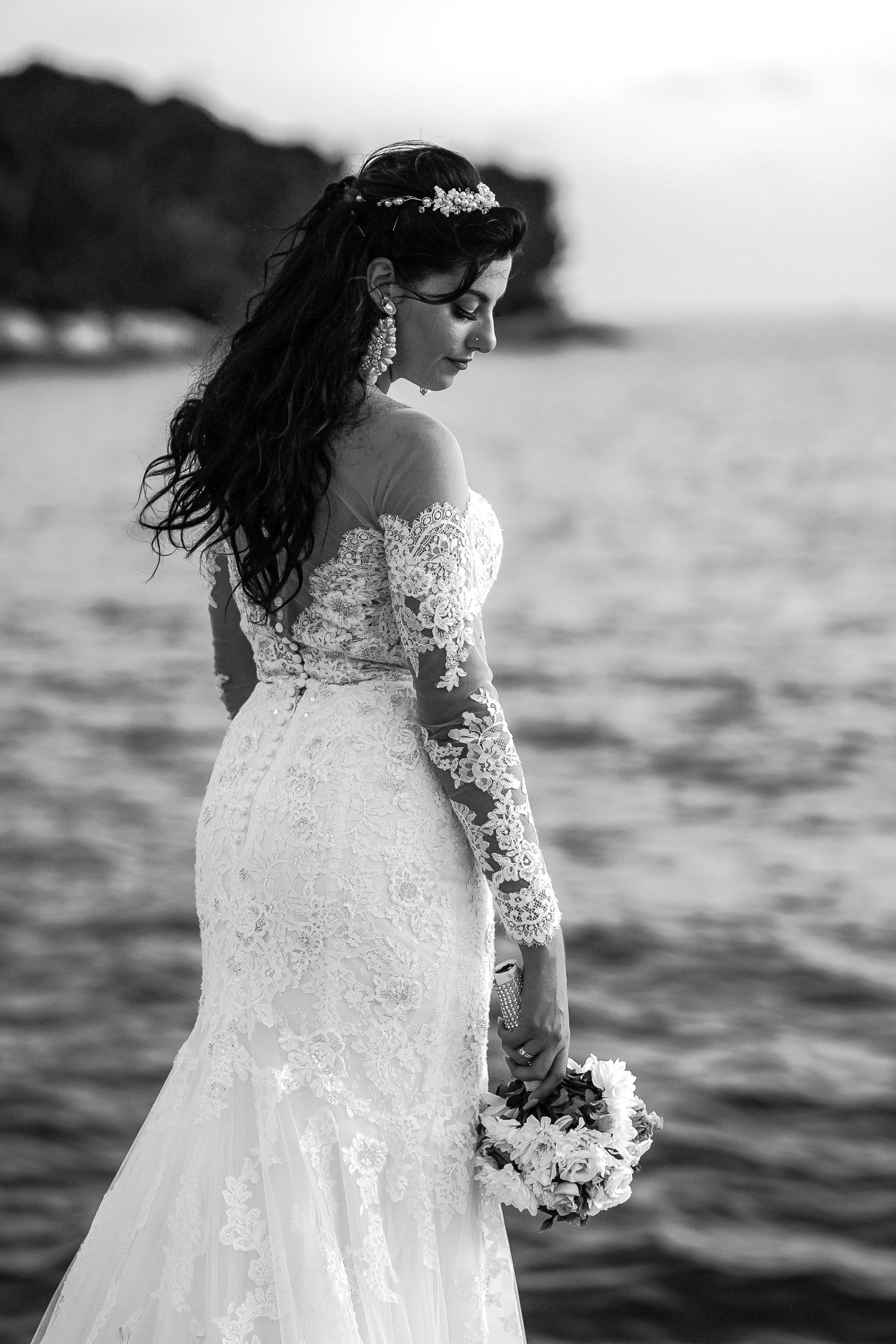 Black and white portrait of a bride in a lace mermaid gown holding a bouquet by the sea.