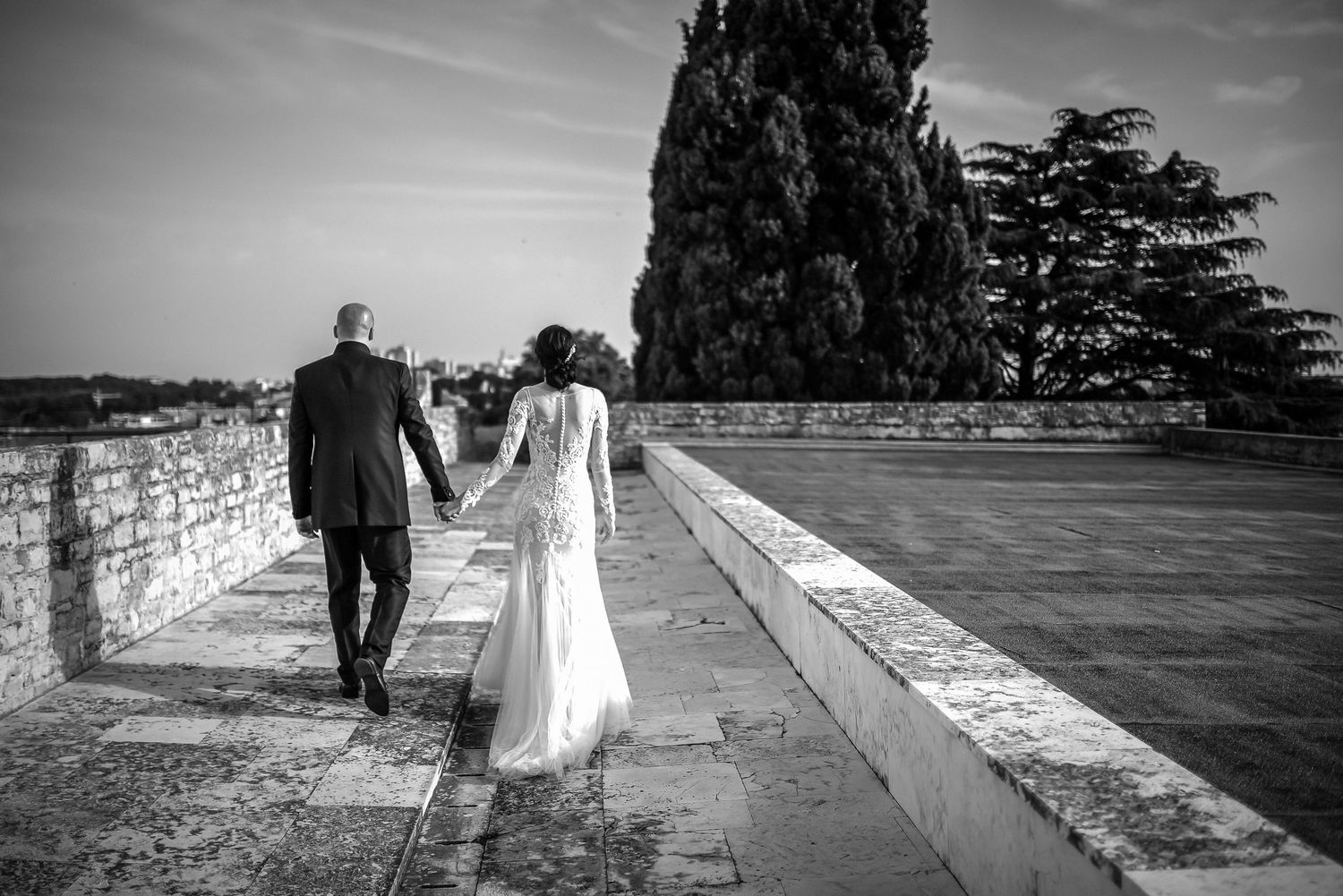 Bride and groom walking hand in hand along a stone wall path in a black and white wedding photo.