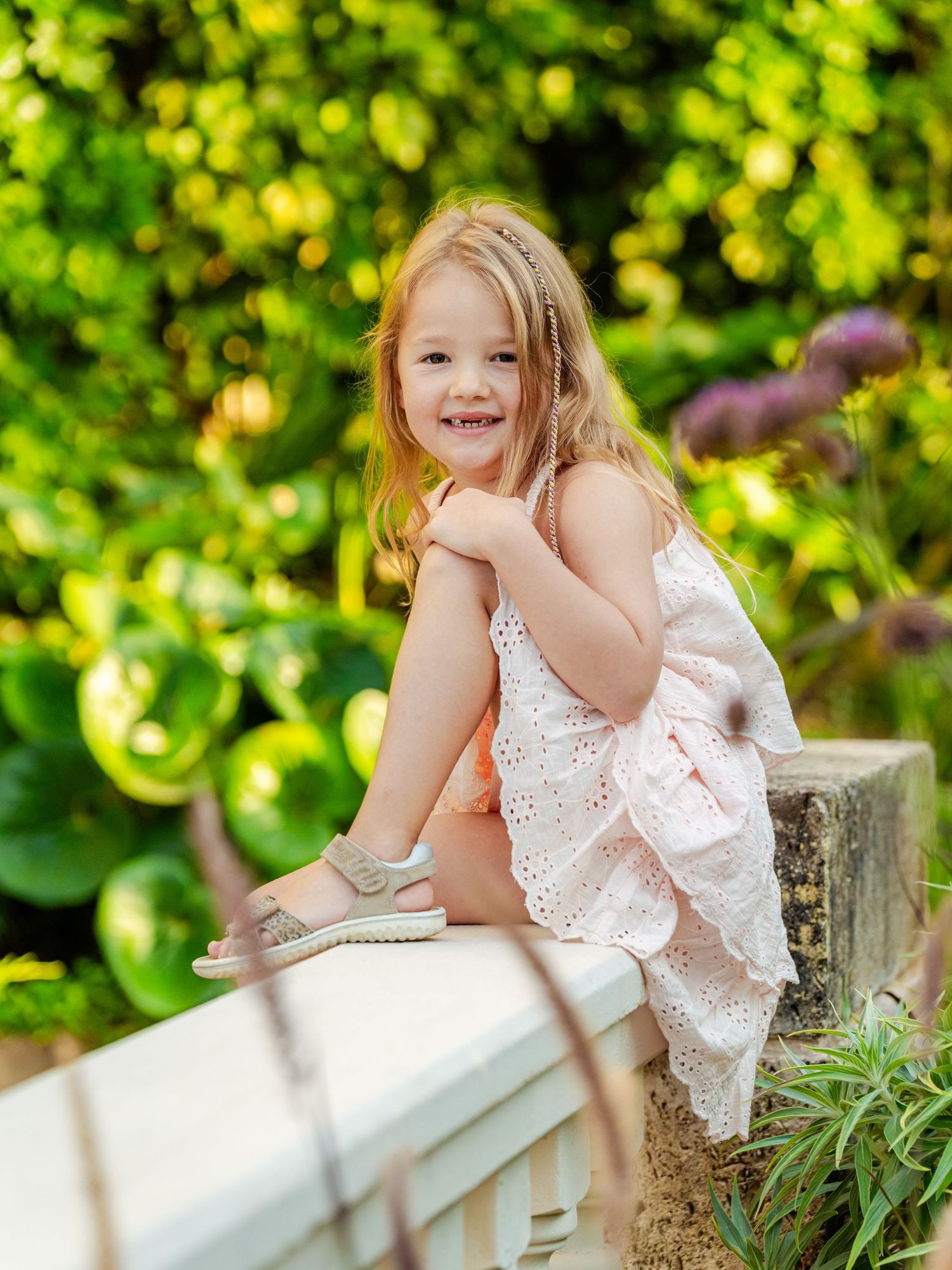 Happy blonde girl in pink dress sitting on stone wall surrounded by lush green garden.