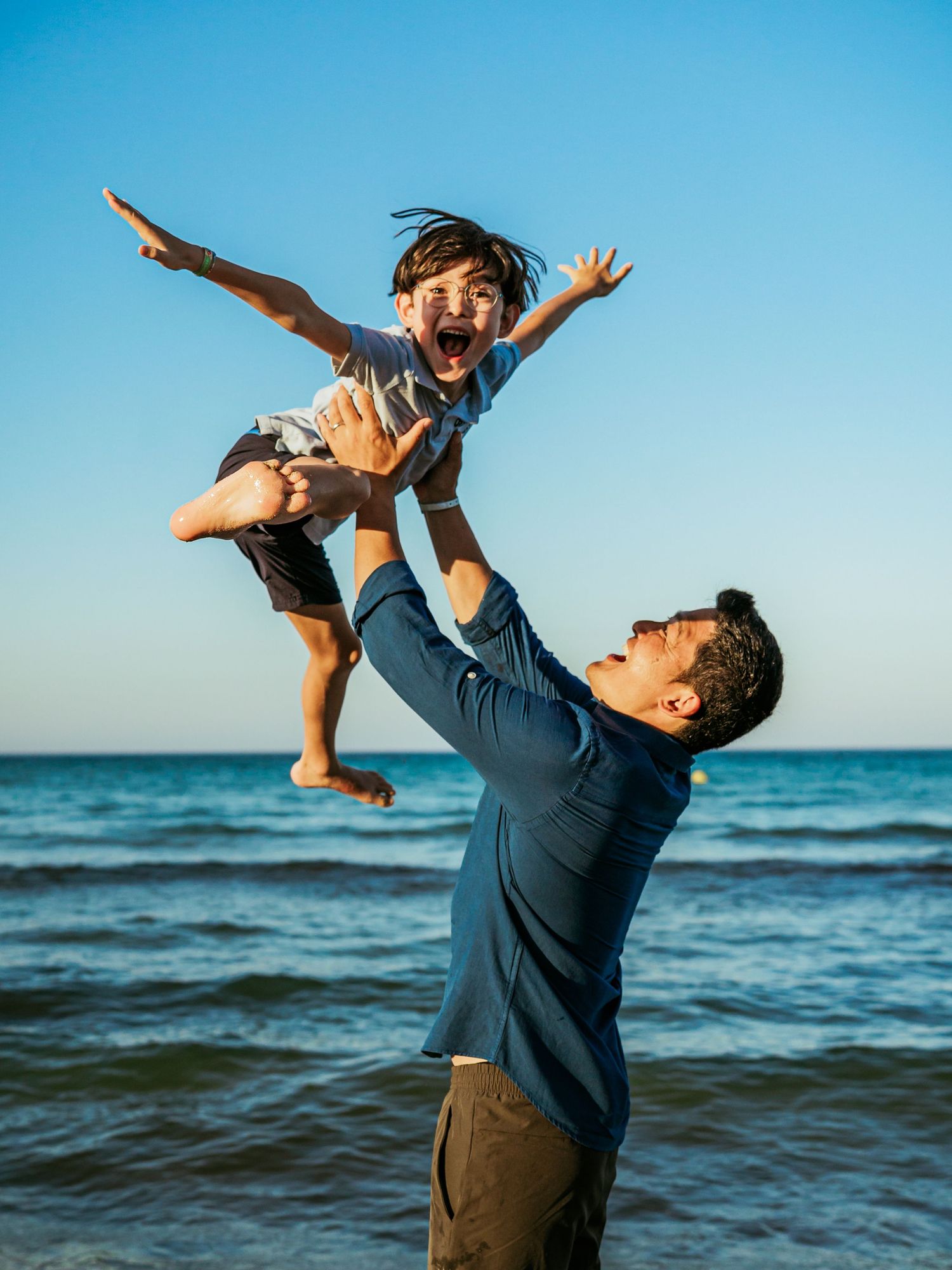Joyful father tossing laughing child high in the air on a sunny beach with blue sky backdrop.