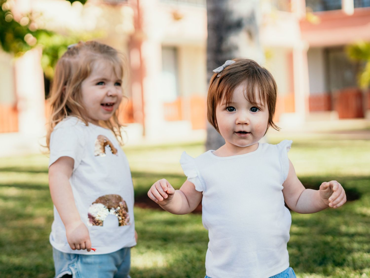 Two cute toddler sisters in white shirts playing together on a sunny green lawn.