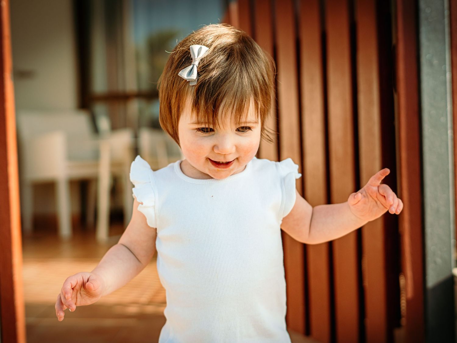 Happy smiling baby girl in white ruffled dress with hair bow standing indoors with arms outstretched.