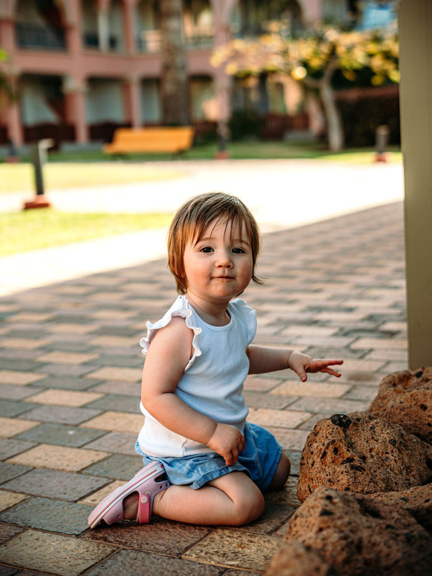 Toddler girl in white top and denim shorts sitting on brick pavement outdoors near decorative rocks.
