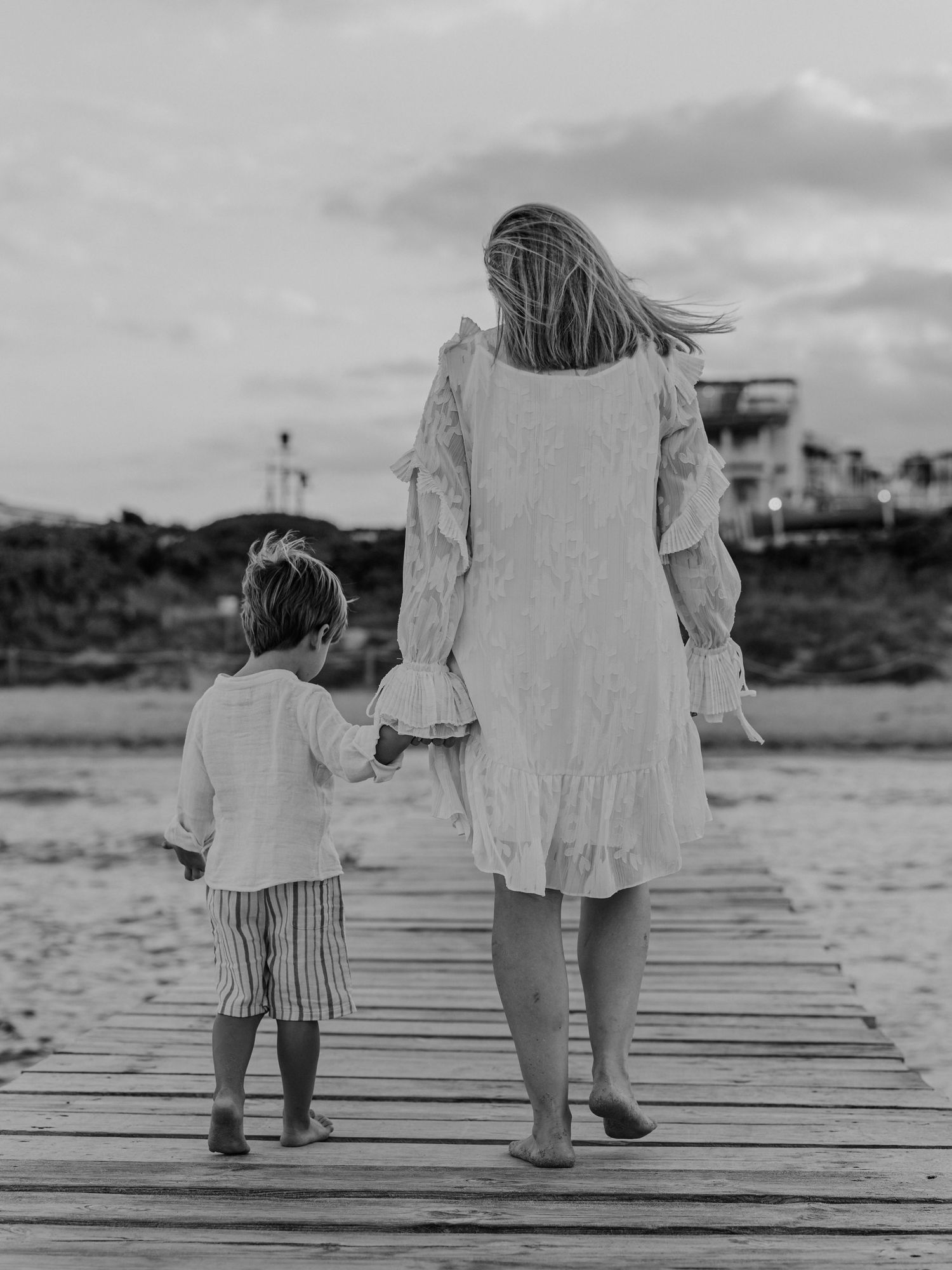 Black and white photo of a mother and young child walking hand in hand along a wooden beach boardwalk.