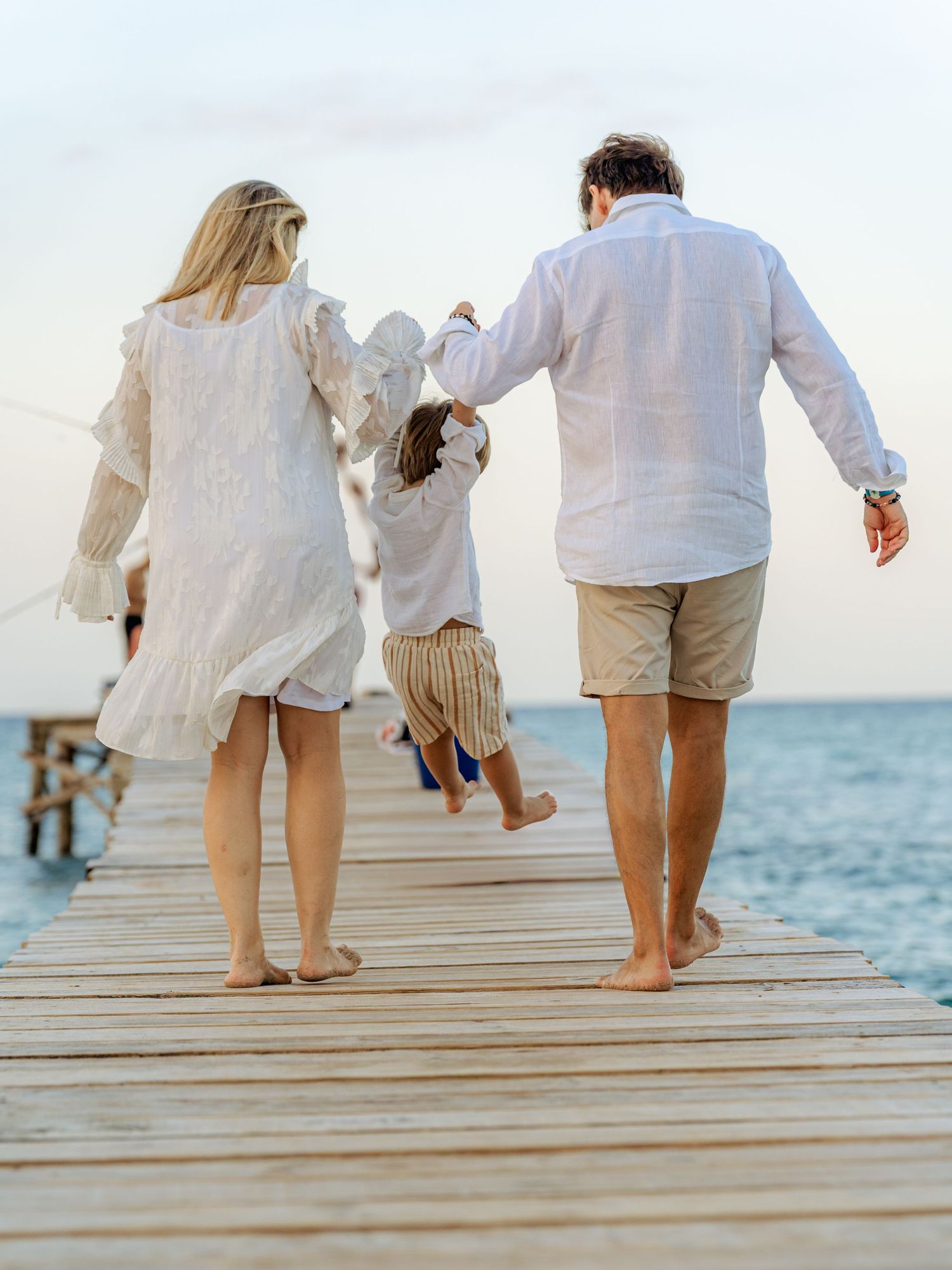 Parents swinging young child between them while walking on seaside dock at dusk near ocean.