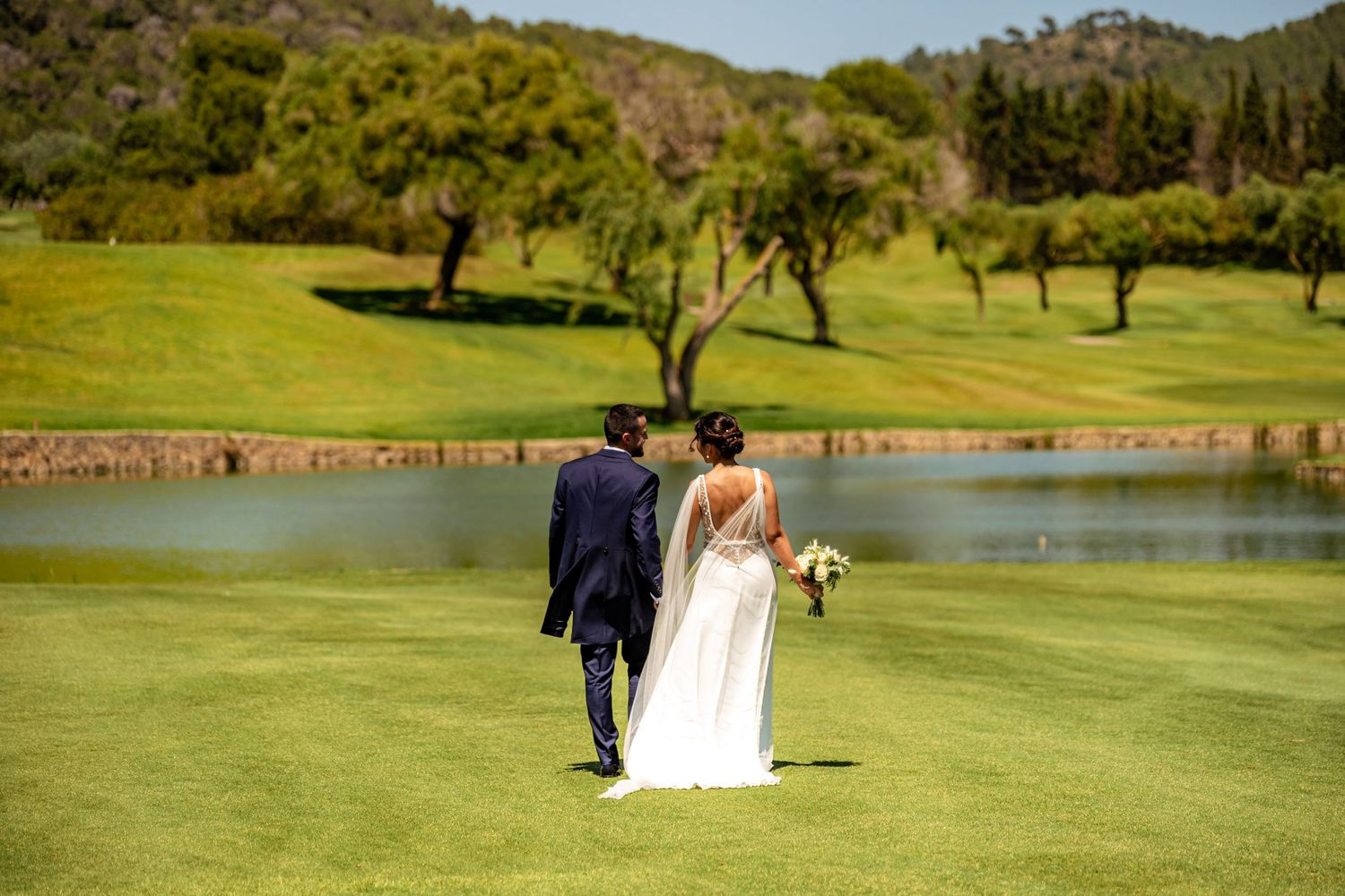 Wedding couple strolls toward serene pond on manicured golf course surrounded by trees.