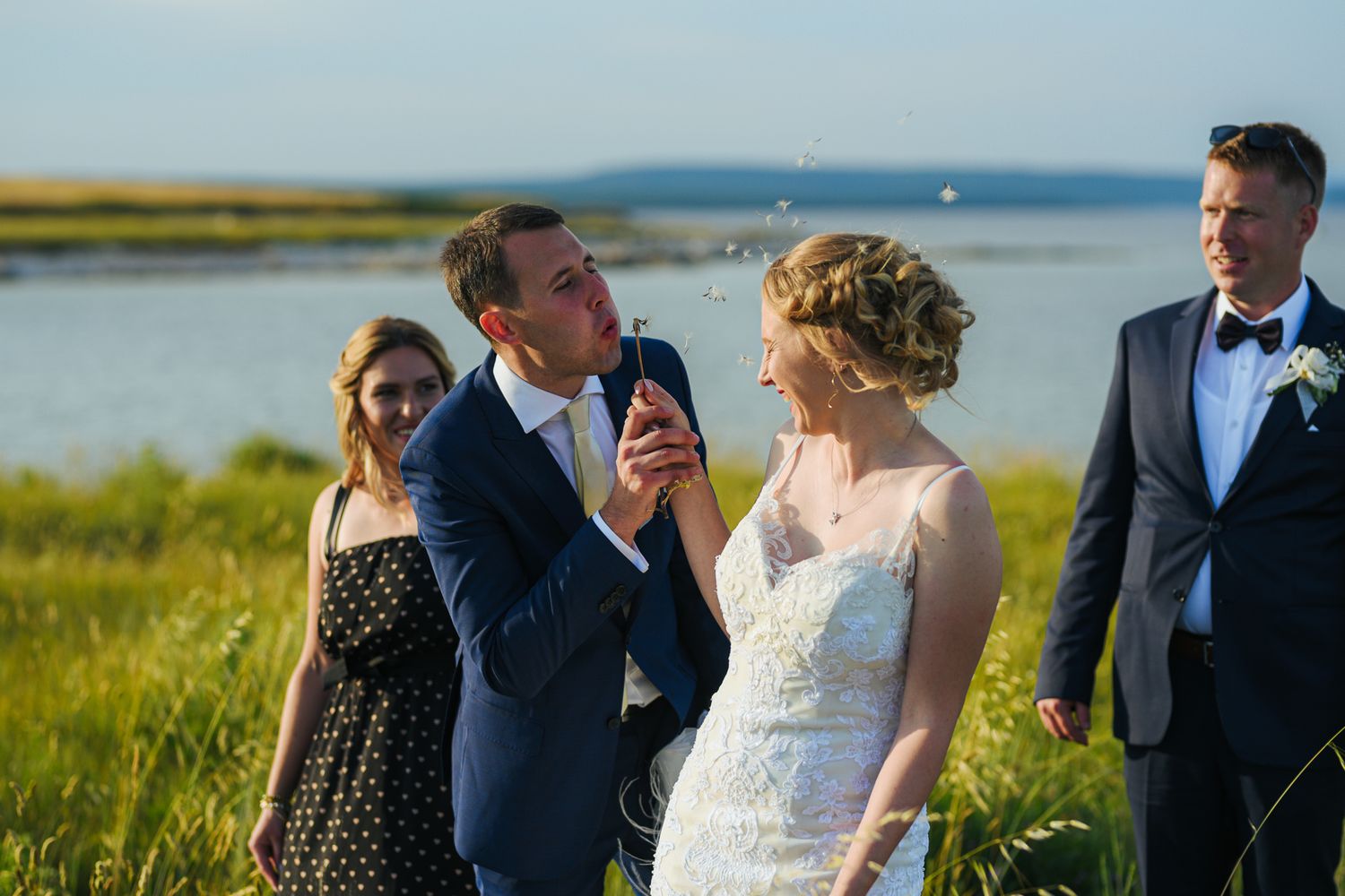 Groom blowing bubbles with bride and guests celebrating in a sunny green coastal field.