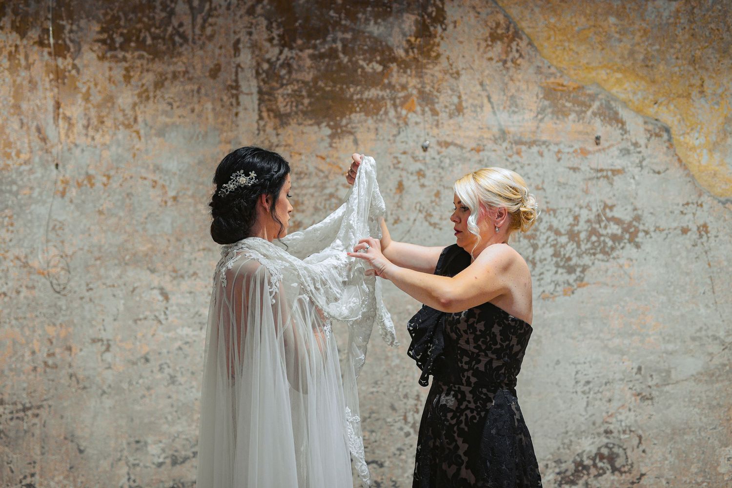 Bride and bridesmaid holding white lace wedding cape against rustic stone wall.