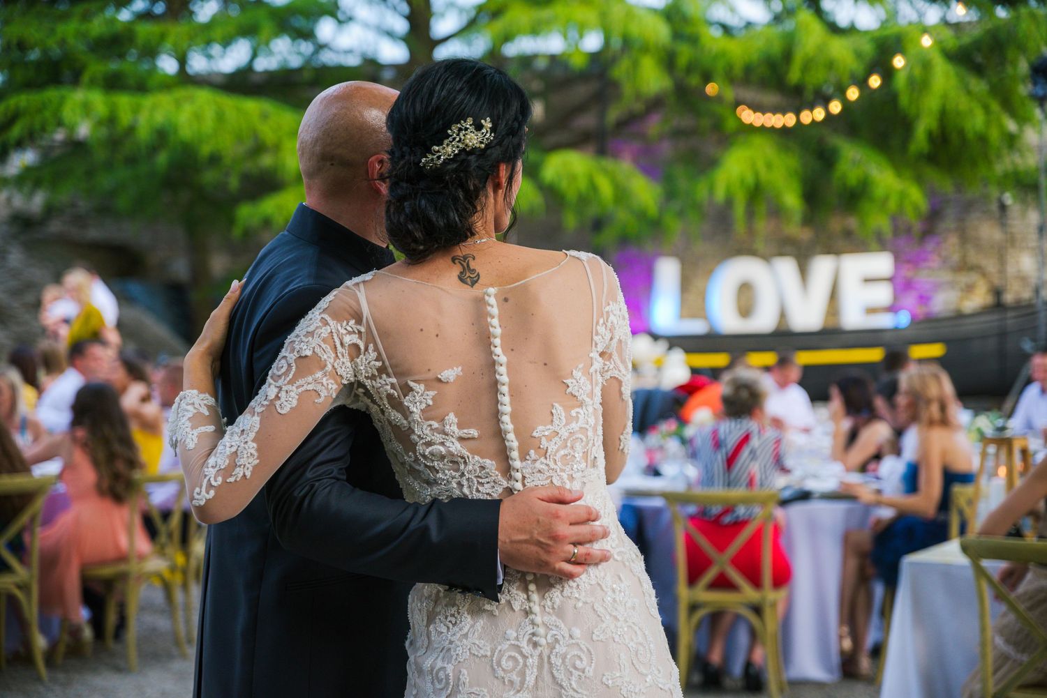 Bride and groom embrace on dance floor with illuminated LOVE sign and guests in background.