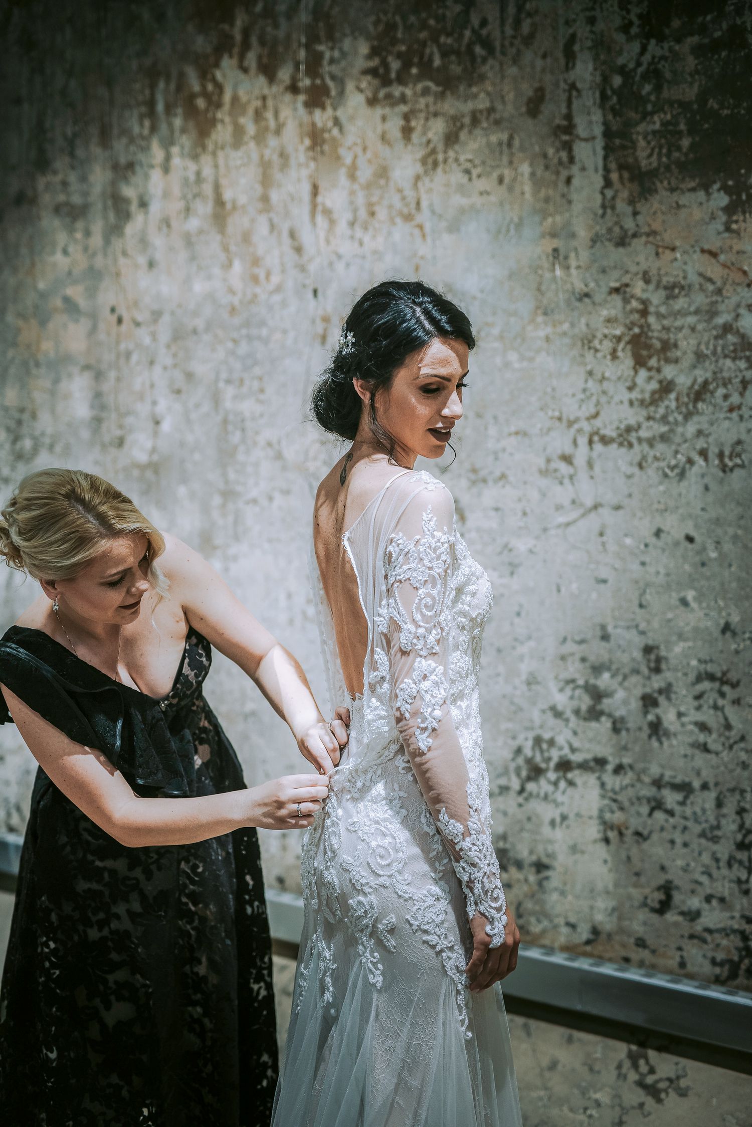 Bride in backless lace wedding gown being buttoned up by woman in black dress.