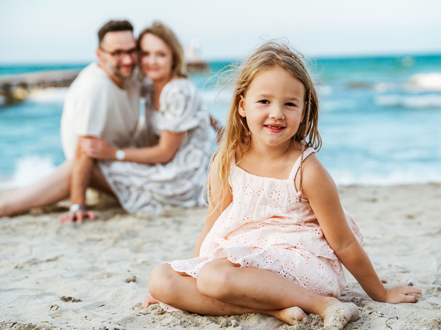 Smiling young girl in pink dress sits on beach while parents relax together behind her.