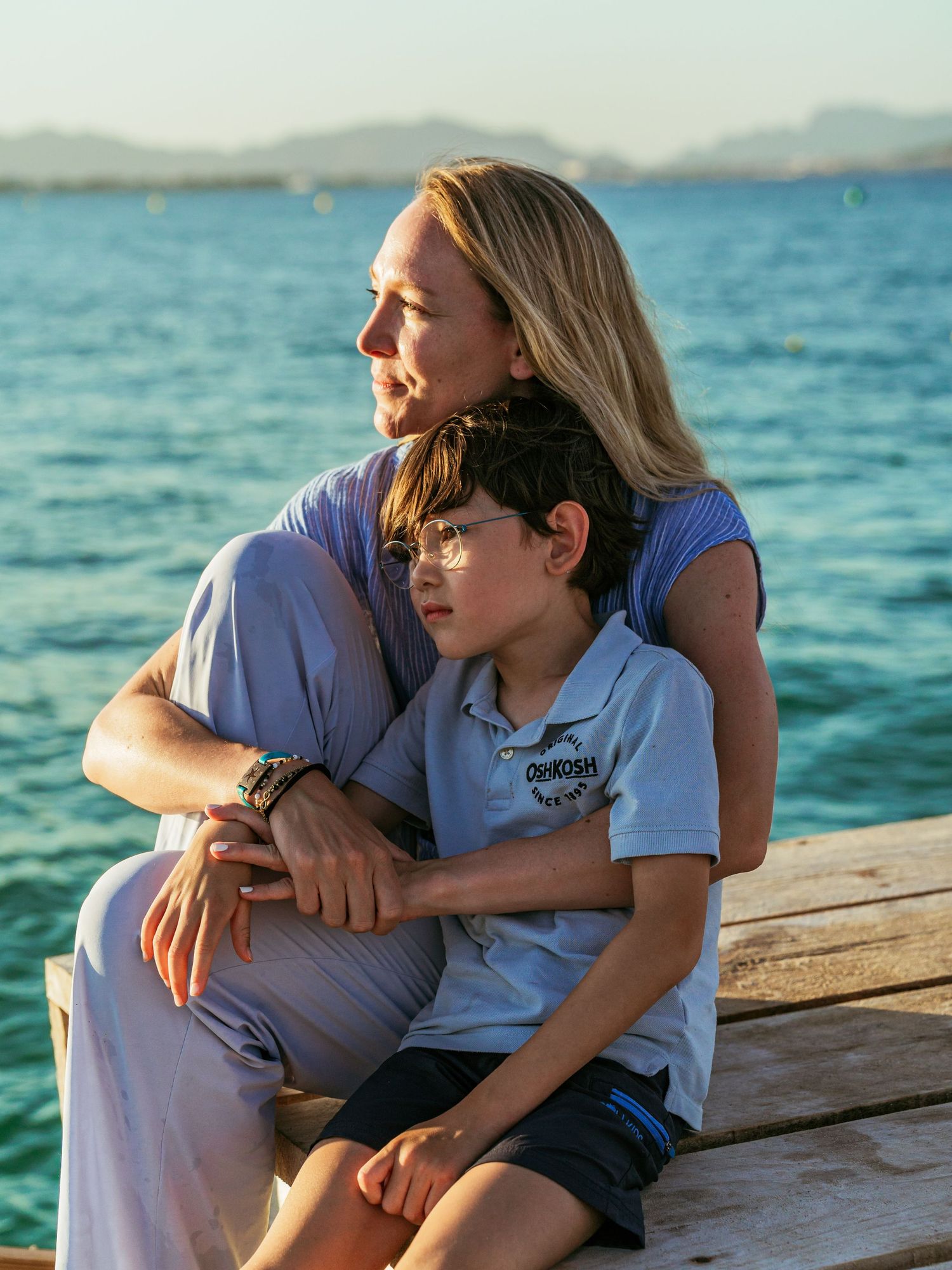 Mother tenderly embracing her young son on a wooden dock with turquoise ocean water in the background.