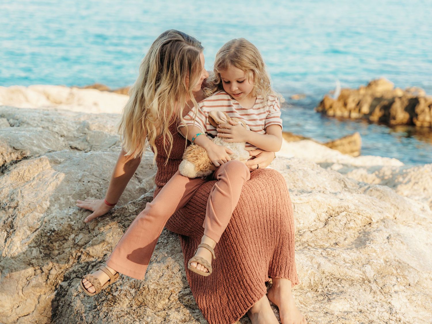 Mother tenderly embracing young child on sun-warmed rocks by the sea.