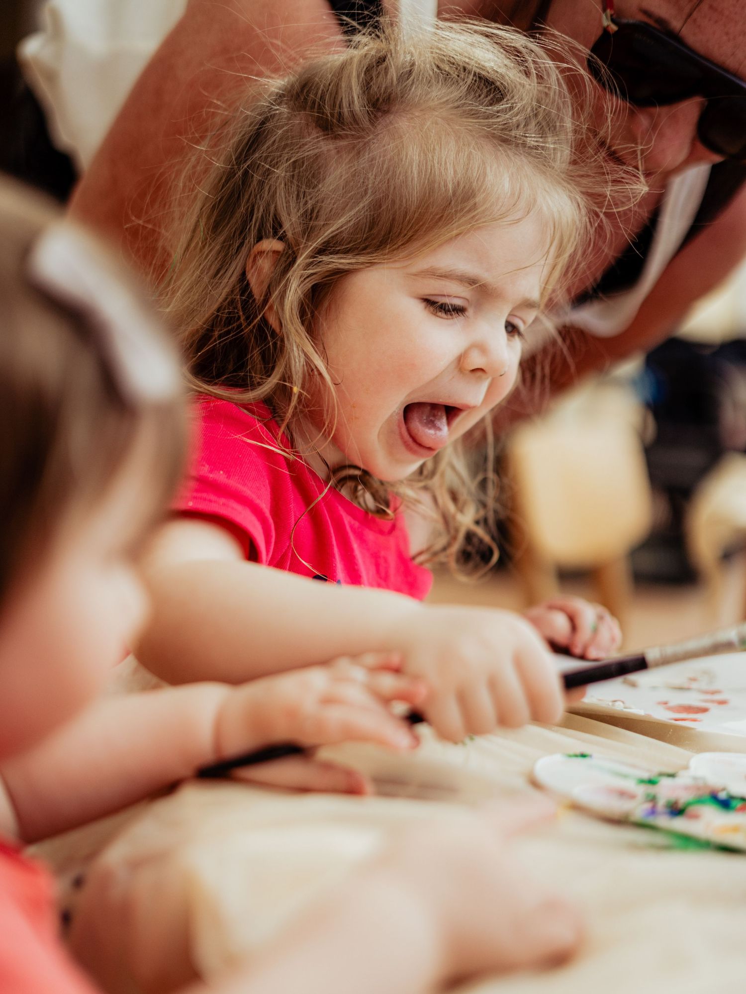 An excited toddler girl in a pink shirt laughs joyfully while painting at a table.
