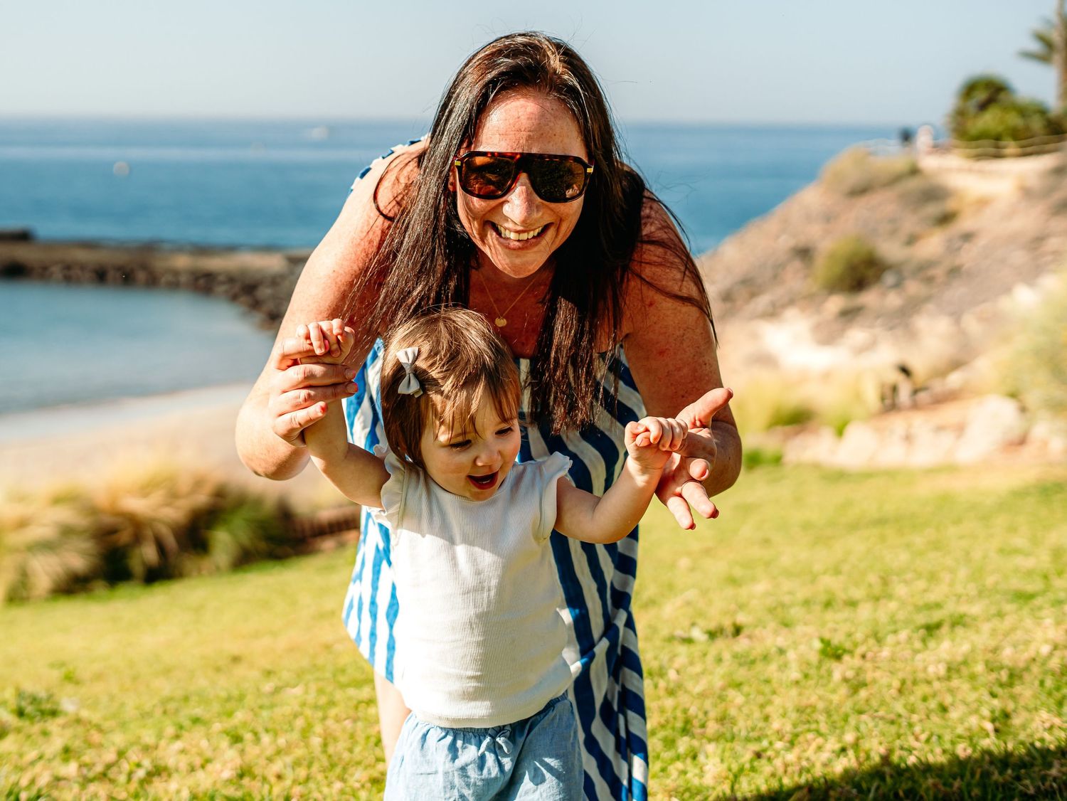 A smiling woman in sunglasses helps a toddler walk on green grass with ocean views in the background.