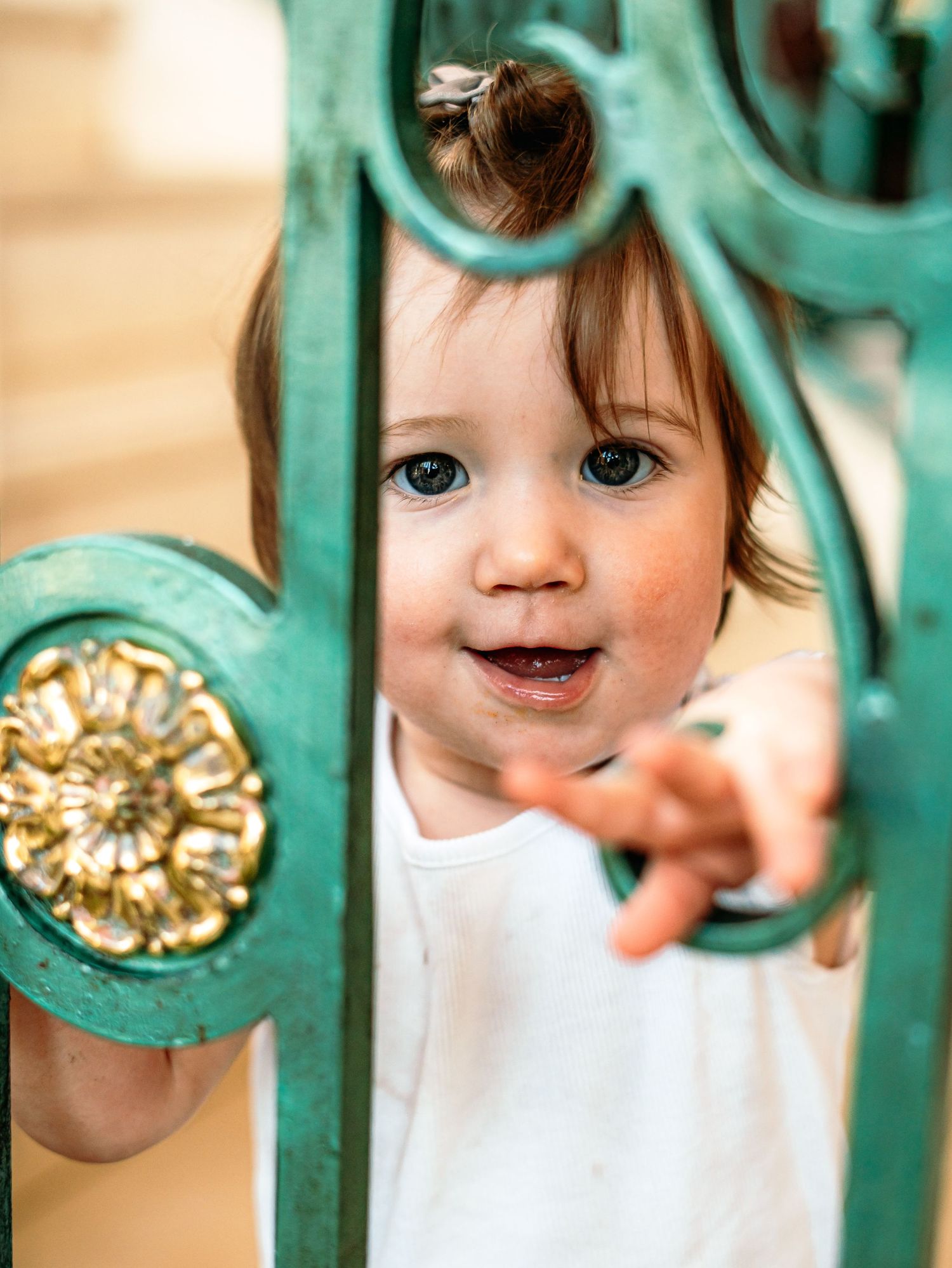 A smiling baby girl reaches through decorative green and gold iron railings toward the camera.