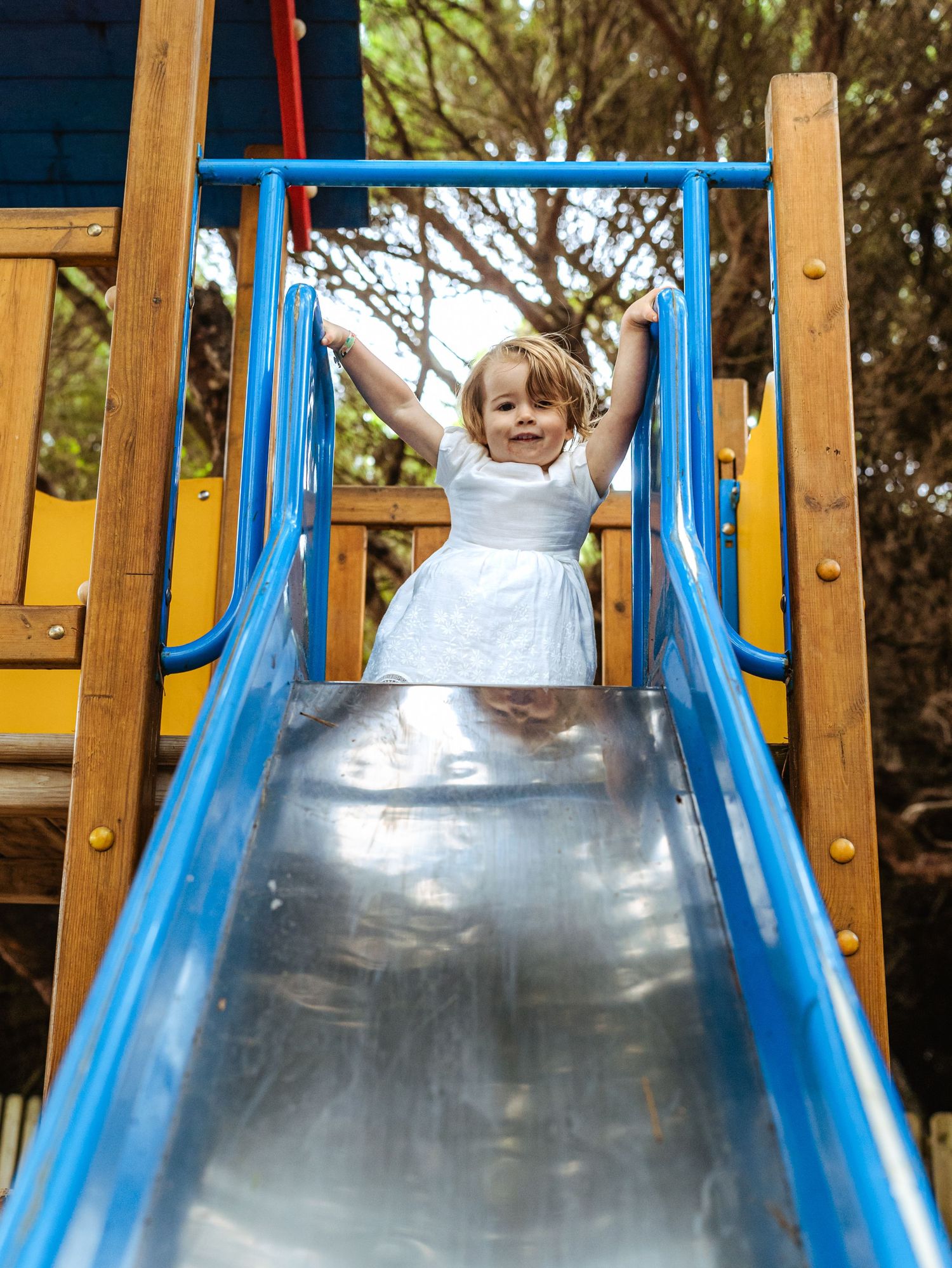 A smiling toddler girl in a white dress rides down a blue playground slide with arms raised.