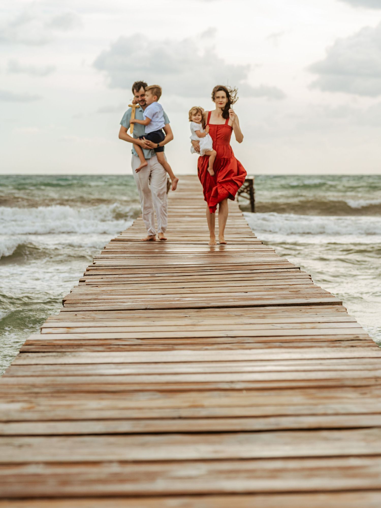 Family strolling on seaside boardwalk, mom in red dress with toddler, dad kissing young son.