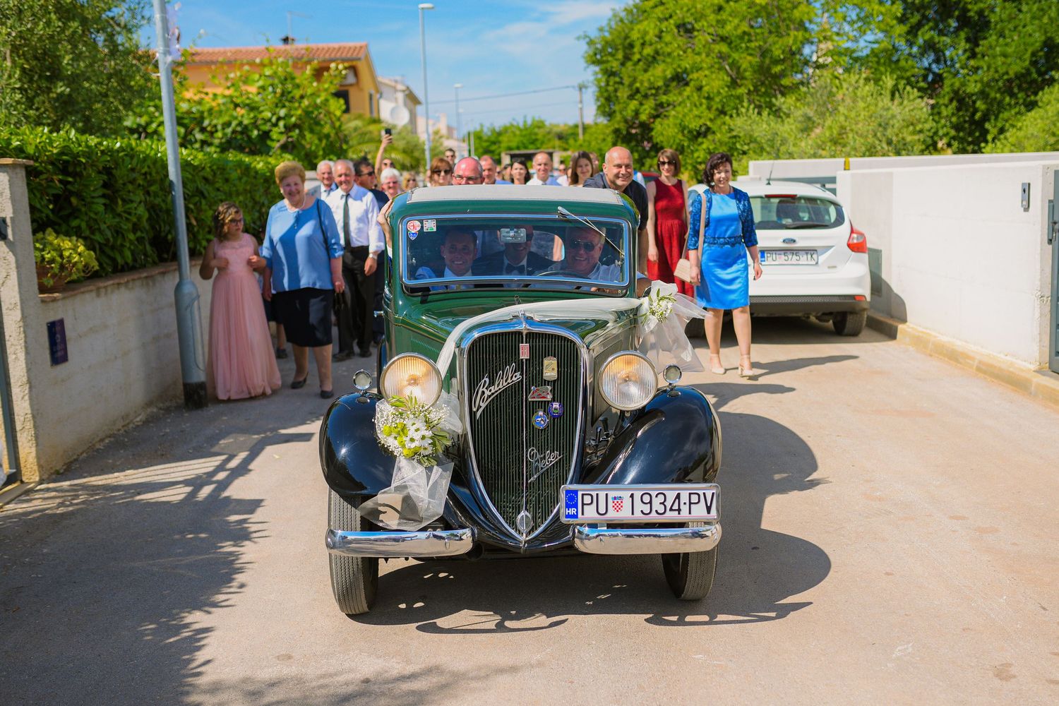 Classic vintage green car decorated with flowers parked outside during outdoor wedding celebration.