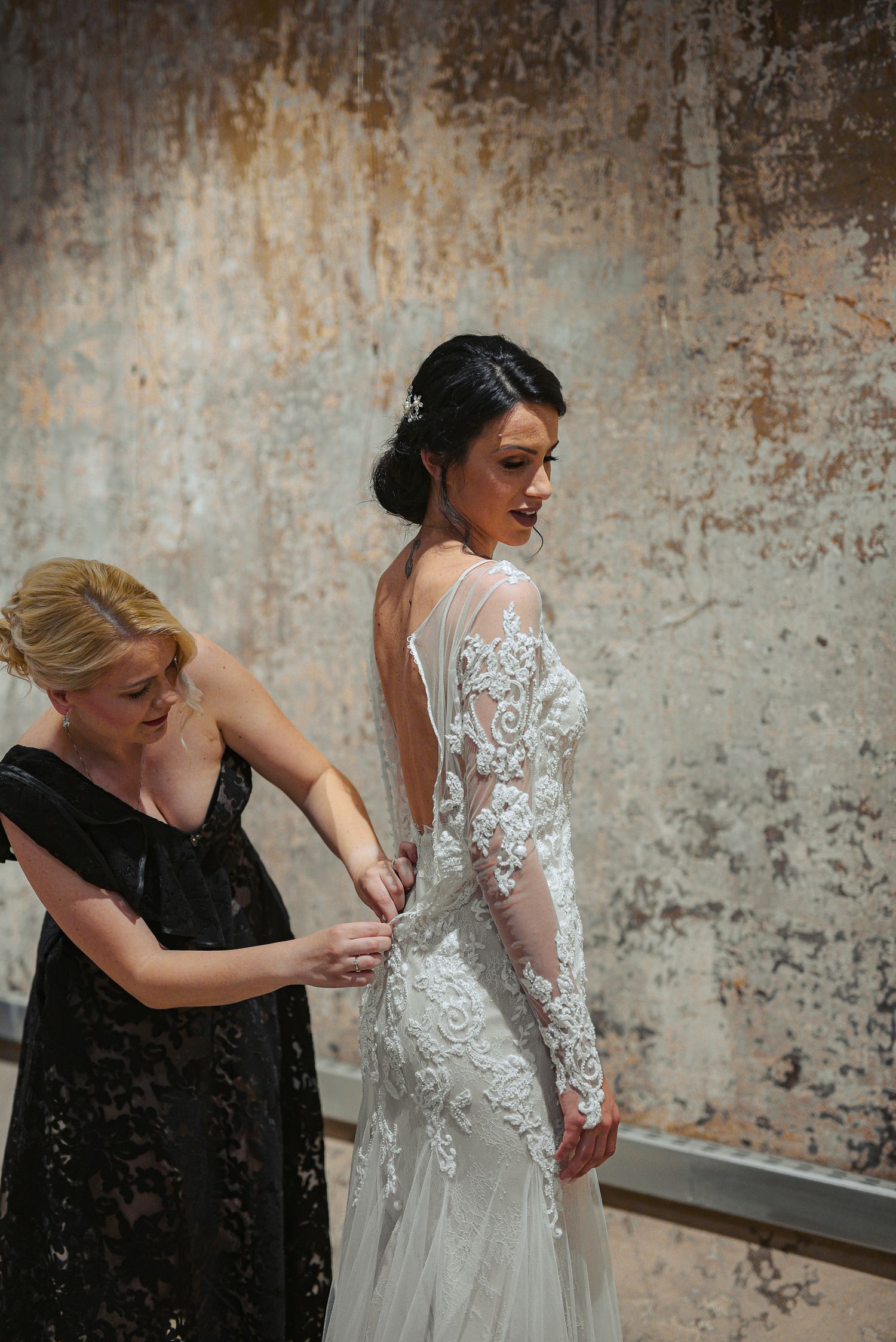 Bride in long sleeve lace wedding gown getting dress buttoned by bridesmaid.