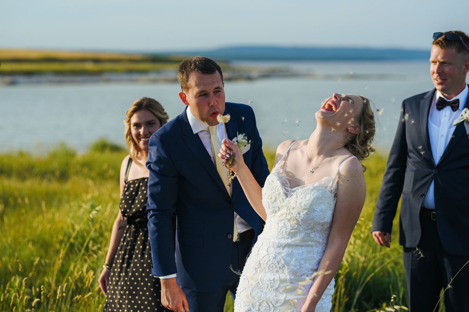 Bride laughing joyfully with groom and wedding guests in a sunny green coastal field.