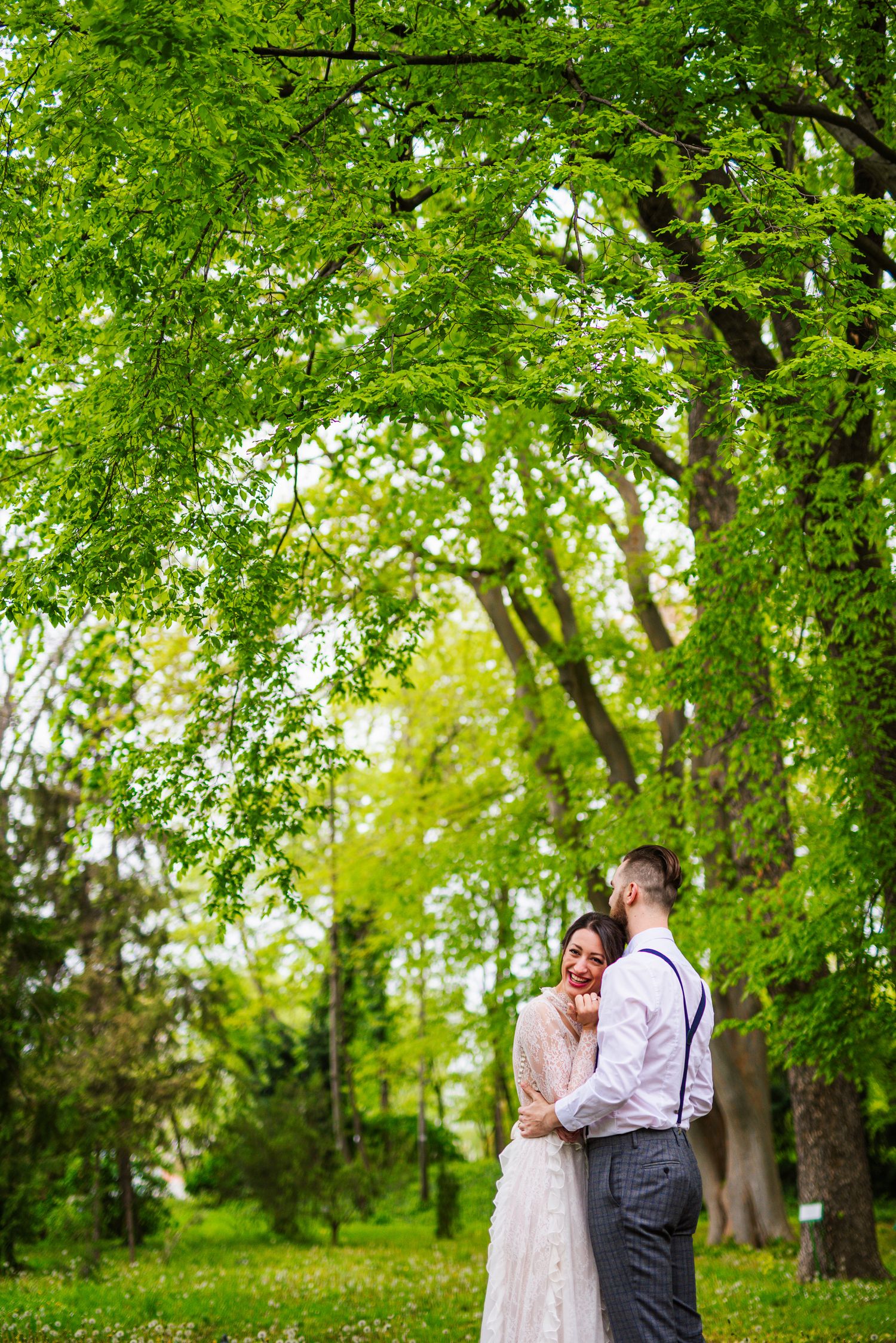 Laughing bride and groom embrace under a canopy of vibrant green trees in a park.