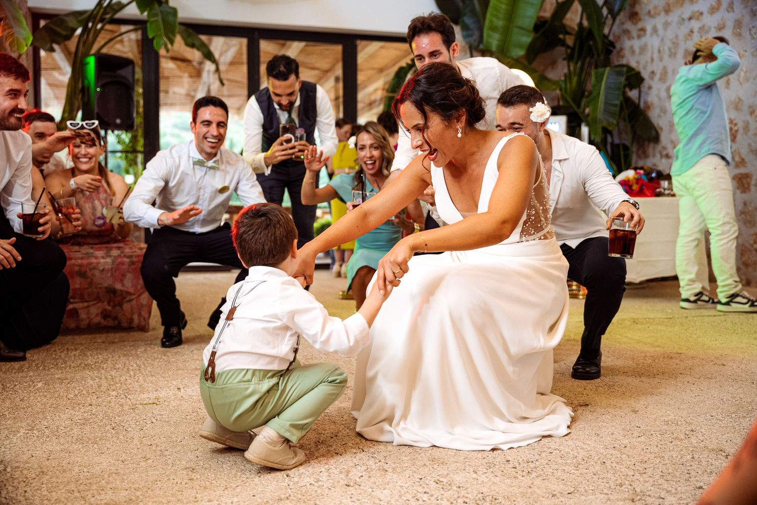 Bride in white gown interacts with young boy on dance floor surrounded by cheering wedding guests.