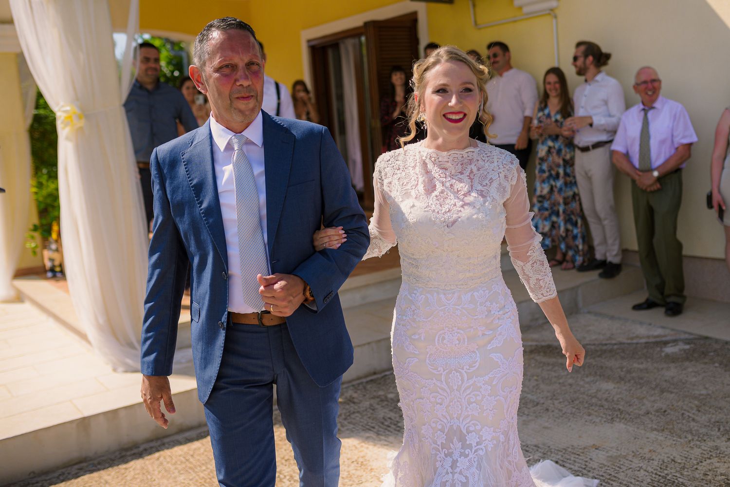 Bride in lace gown walking arm-in-arm with man in blue suit at sunny outdoor wedding venue.