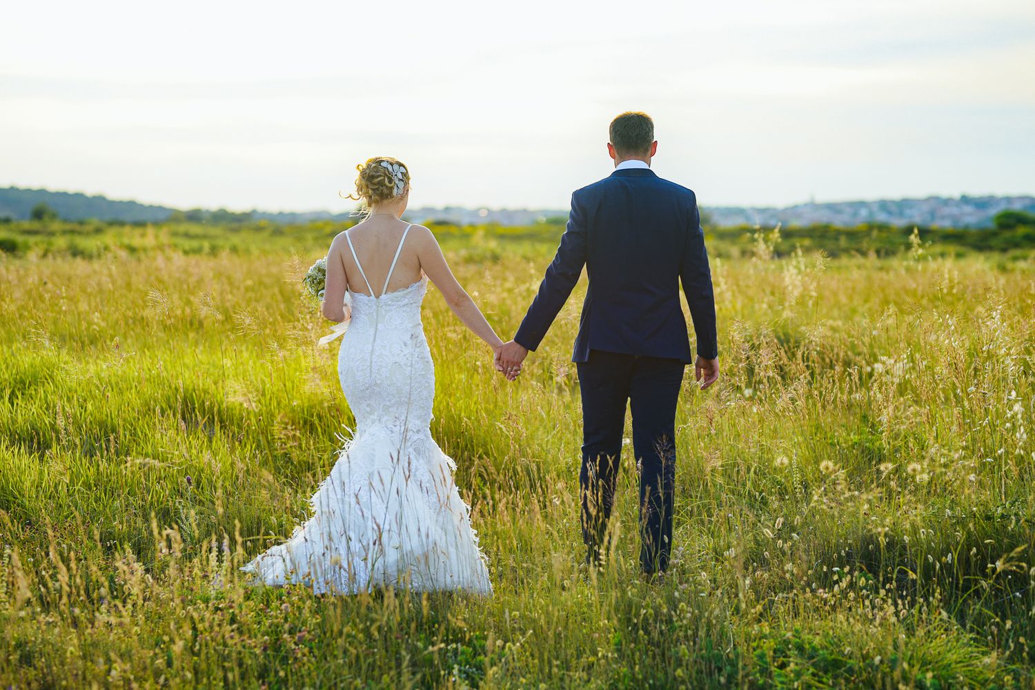 Bride and groom walking hand in hand through lush green meadow on their wedding day.
