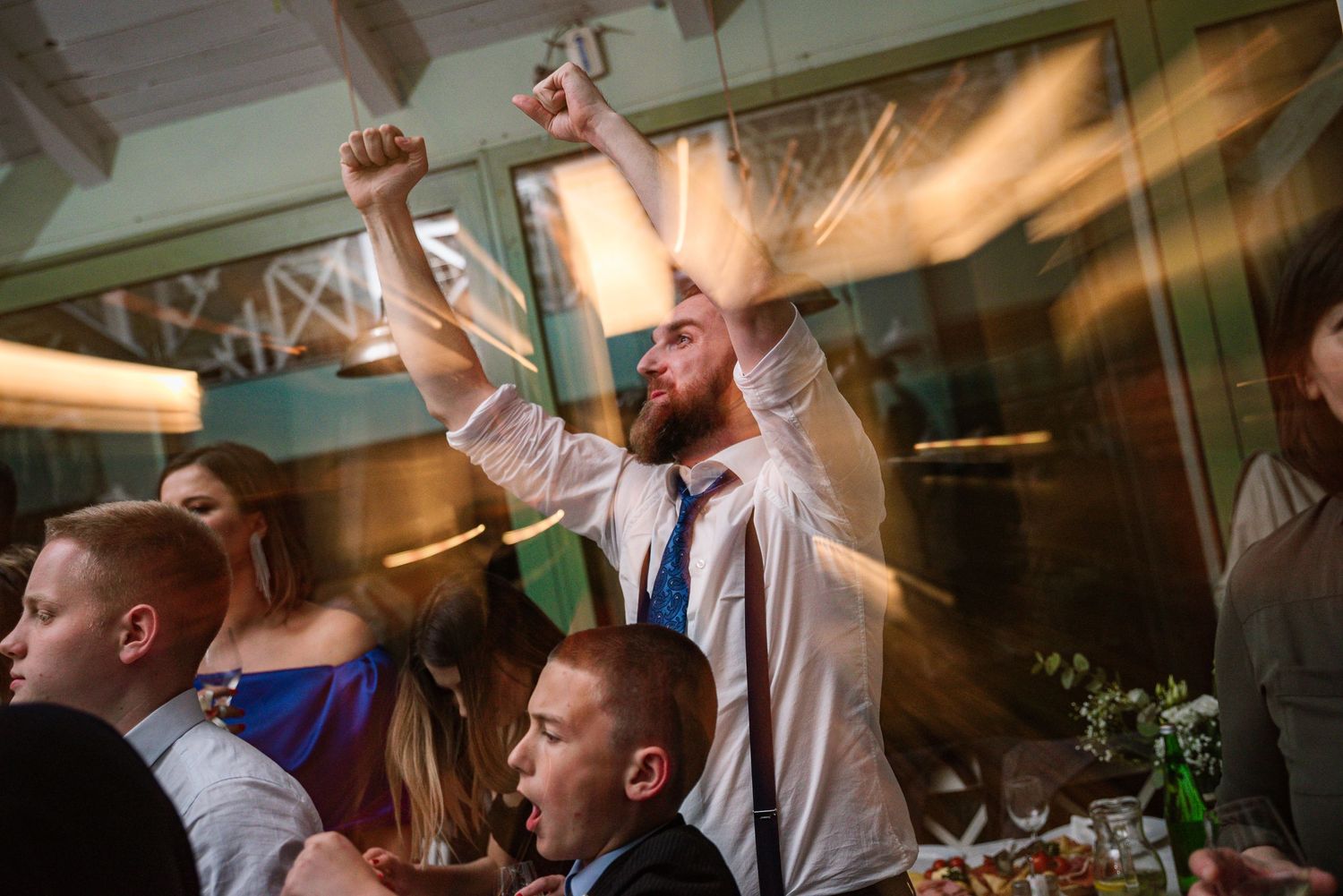 Excited bearded man raises fists in celebration at a wedding reception dinner party.