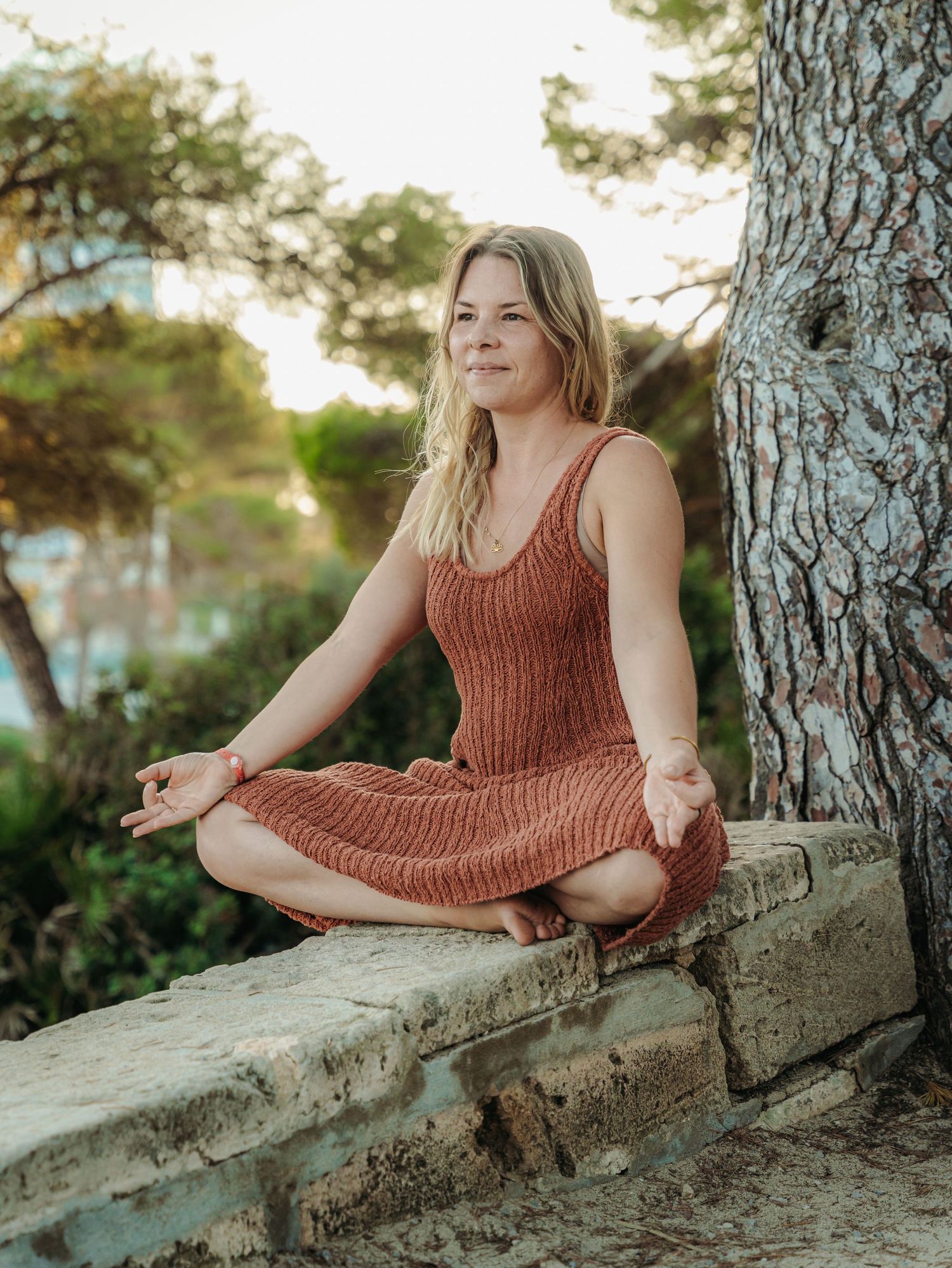 Woman in rust knit dress meditating in lotus pose on stone wall outdoors near tree.