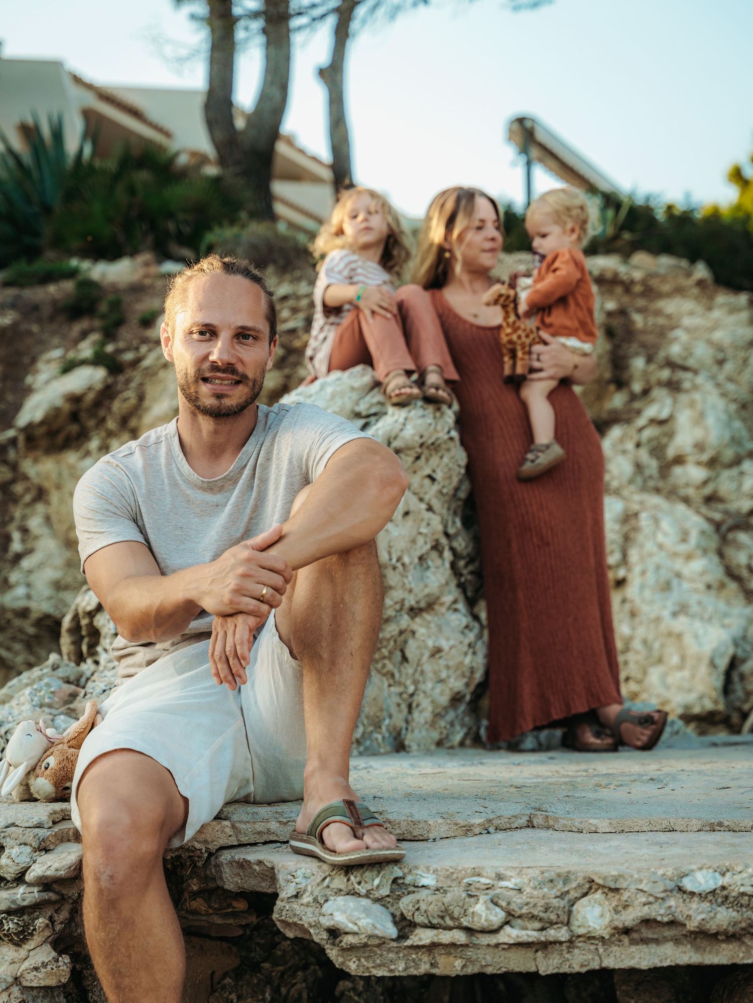 A man sits on stone steps while a woman holds two children among rocks and trees in warm evening light.
