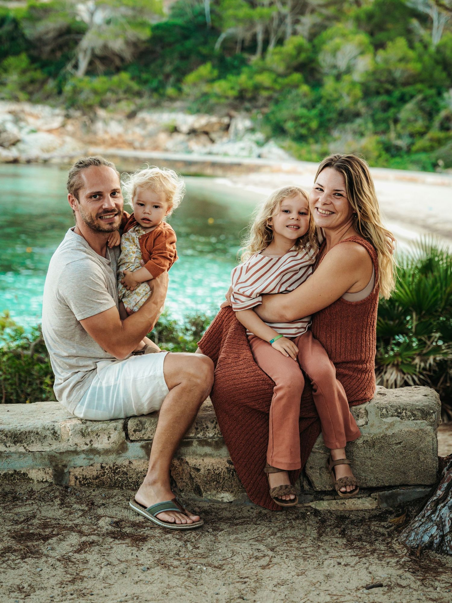 Smiling family with two young children sitting on stone wall with beautiful blue cove behind them.
