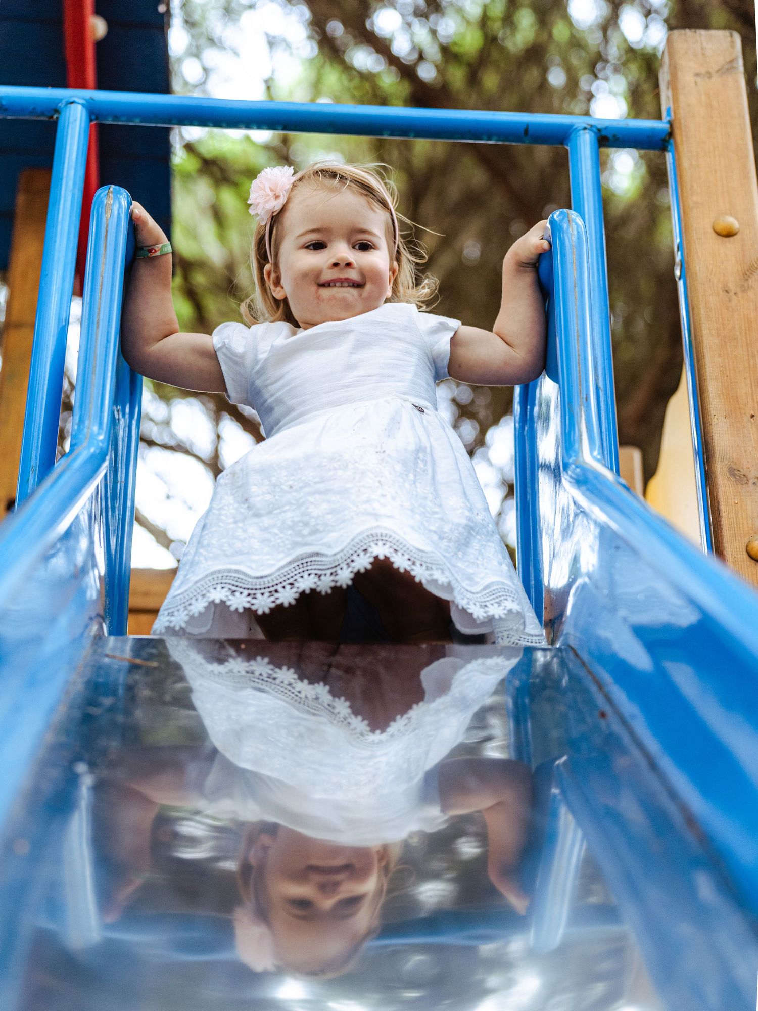 Cheerful little girl in white dress smiling while sitting at top of a blue playground slide.