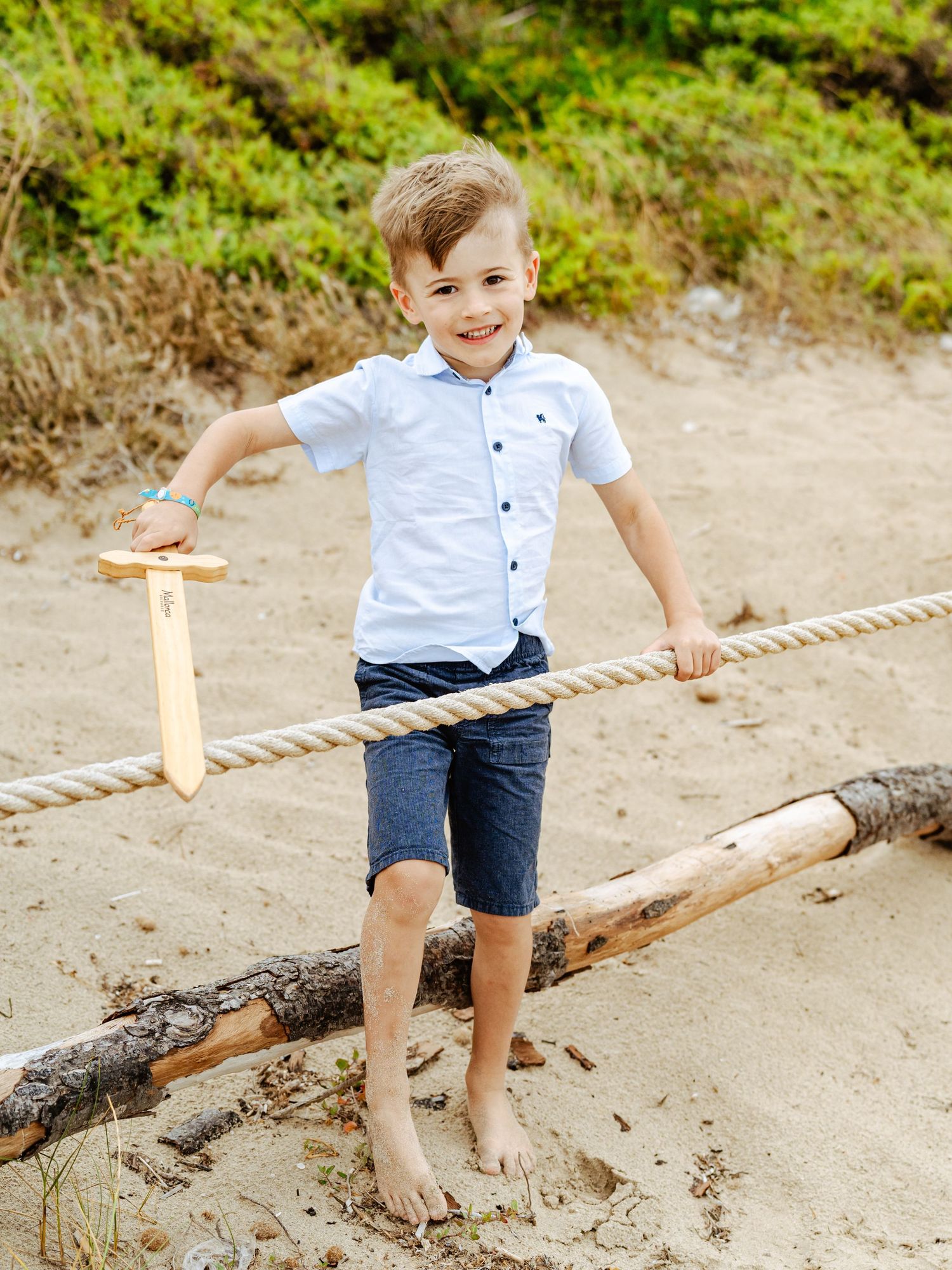 A smiling boy in a white shirt and navy shorts holds a wooden sword while balancing on a log rope at the beach.