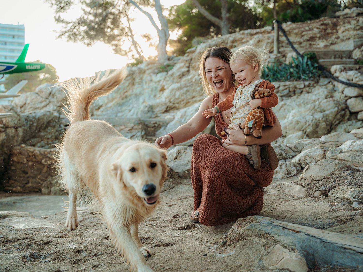 Laughing mother sits with toddler and stuffed giraffe as a golden retriever walks nearby outdoors.