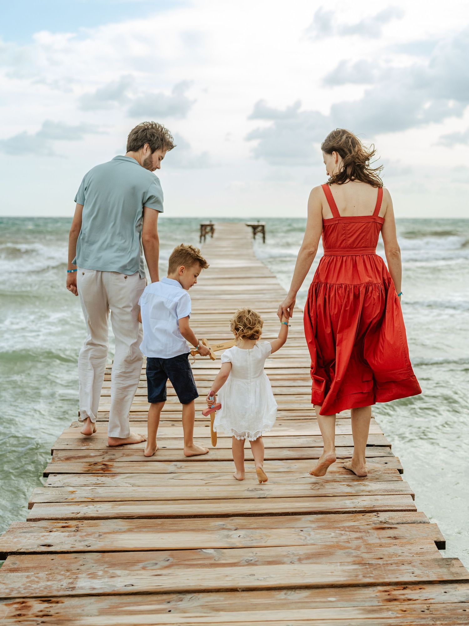 Family of four holding hands walking on wooden pier toward ocean on cloudy day.