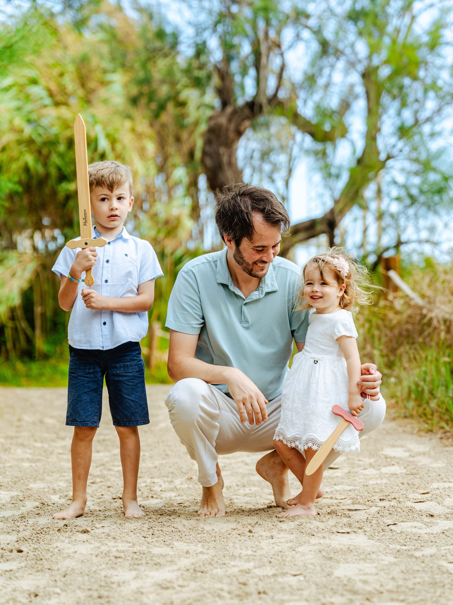 Father crouching on sandy beach path with young son holding toy sword and cheerful toddler daughter.