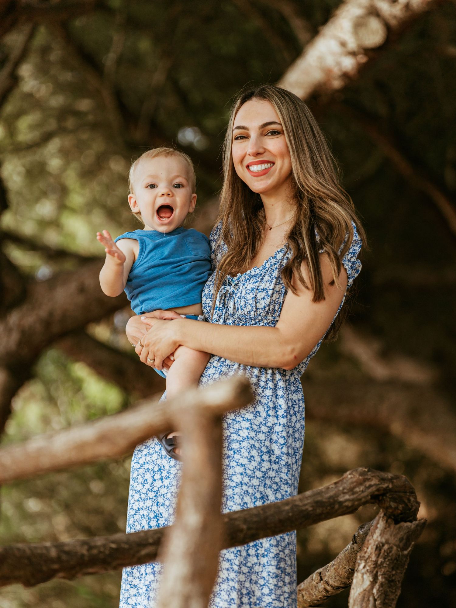Smiling mother in a floral dress holding her expressive baby boy outdoors among trees.
