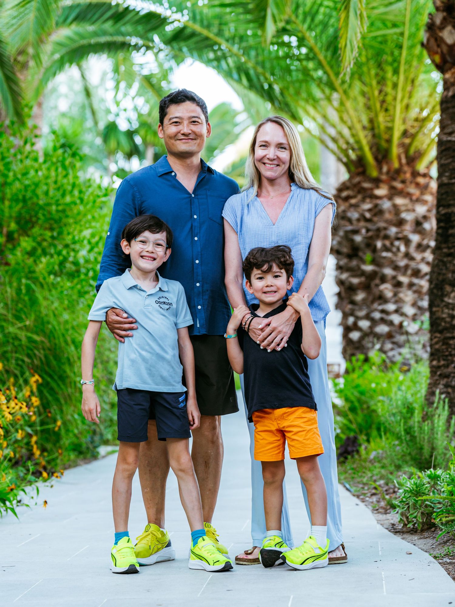 Happy family of four with two boys posing on tropical palm tree lined pathway.