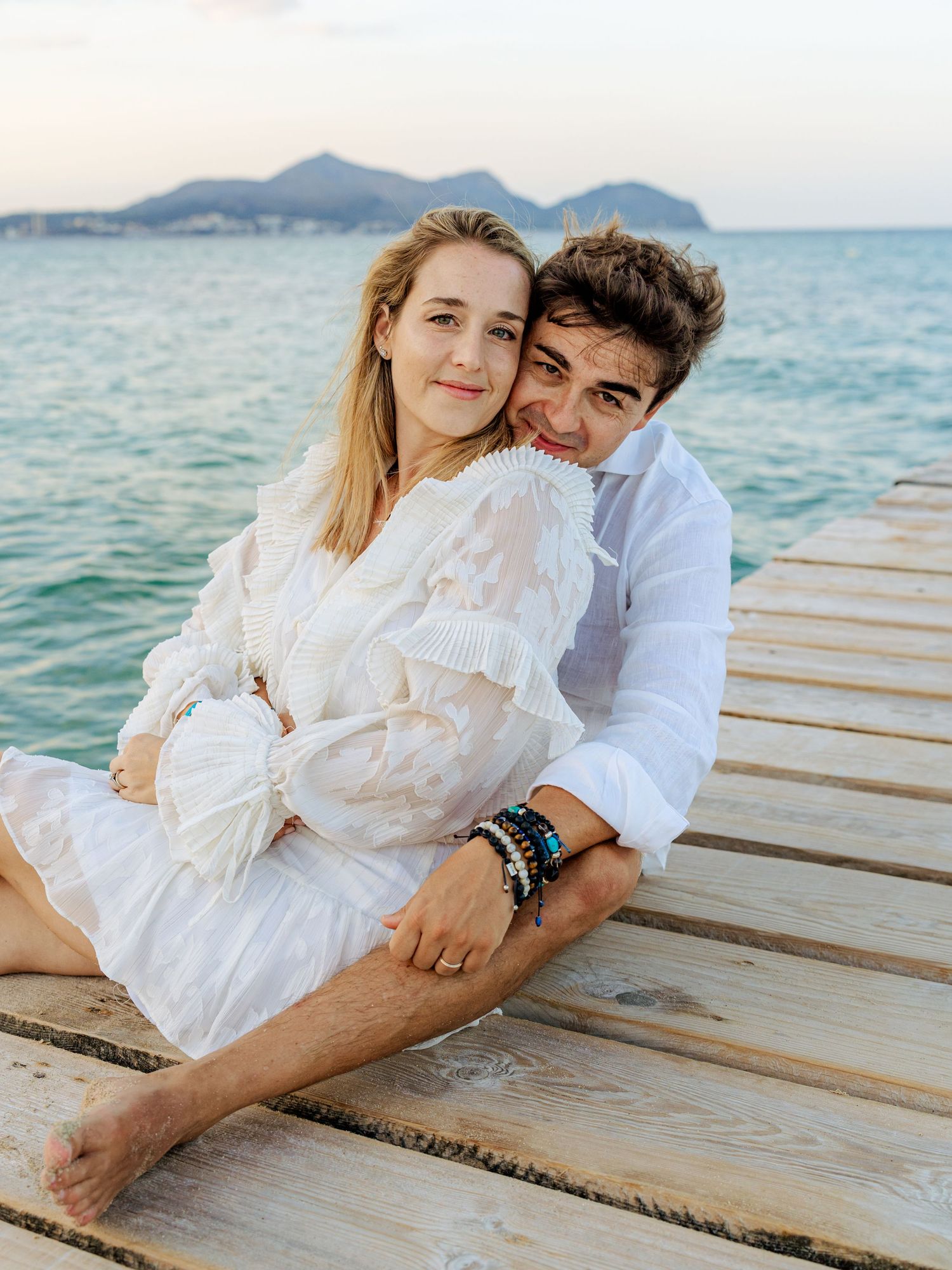 Smiling couple sitting closely together on a wooden pier with turquoise ocean and mountains behind them.