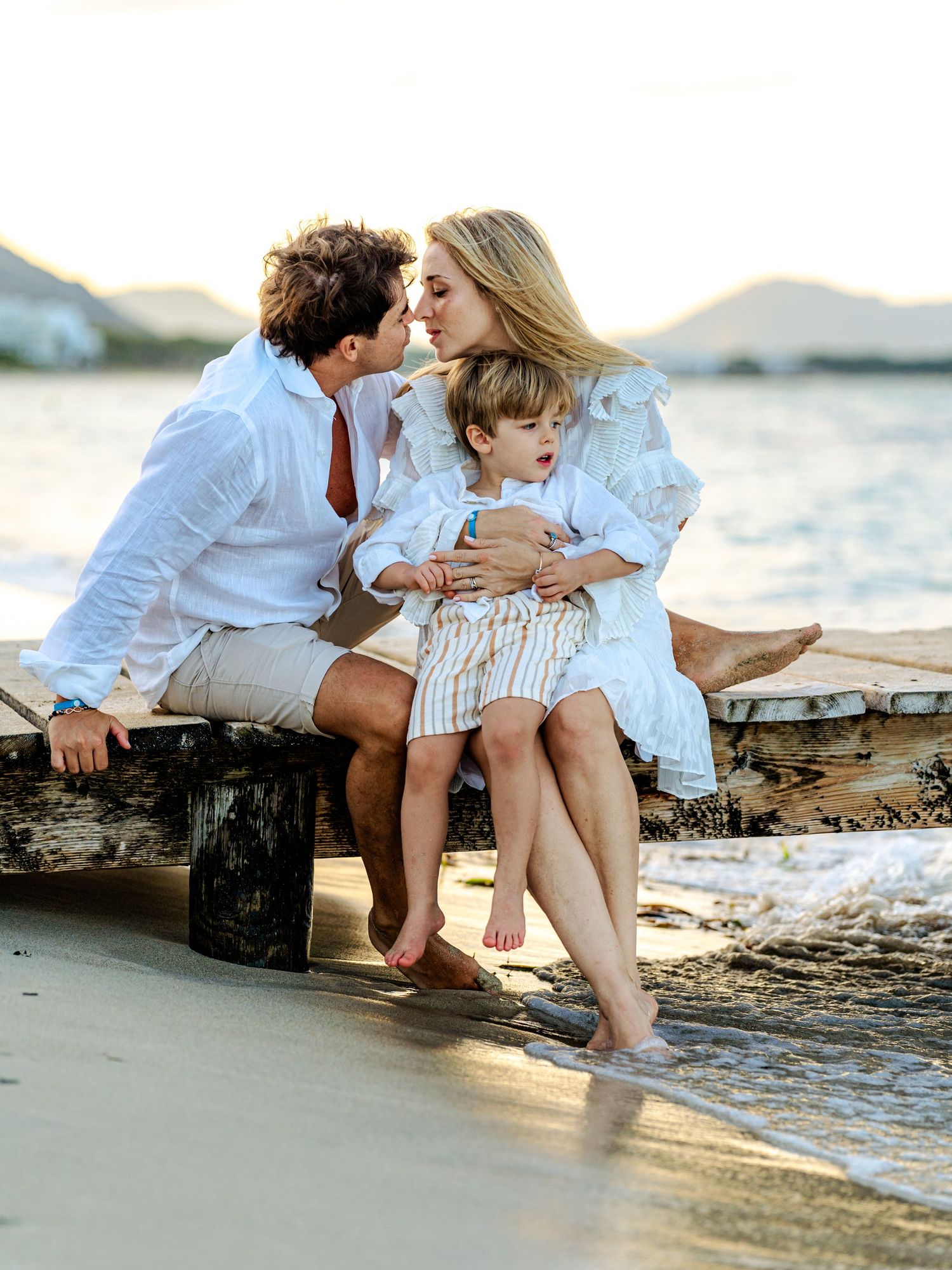 Family of three sharing a tender moment on a beach dock at sunset, parents kissing over their toddler.