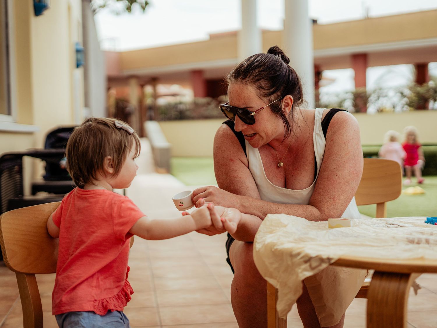 A mother hands something to her young toddler at an outdoor wooden table on a sunny day.