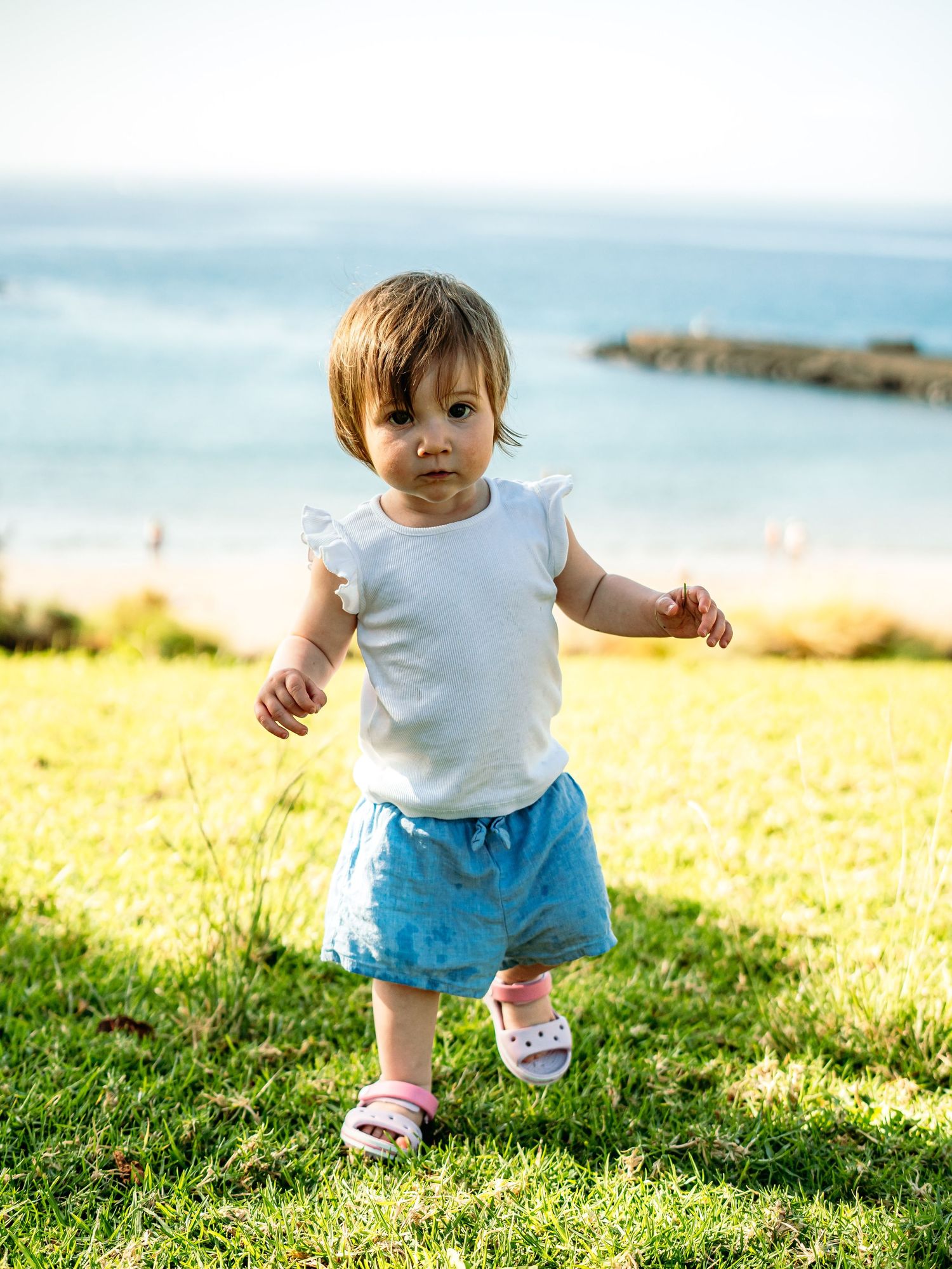 Baby girl in white top and blue shorts walking on green grass with ocean view behind her.